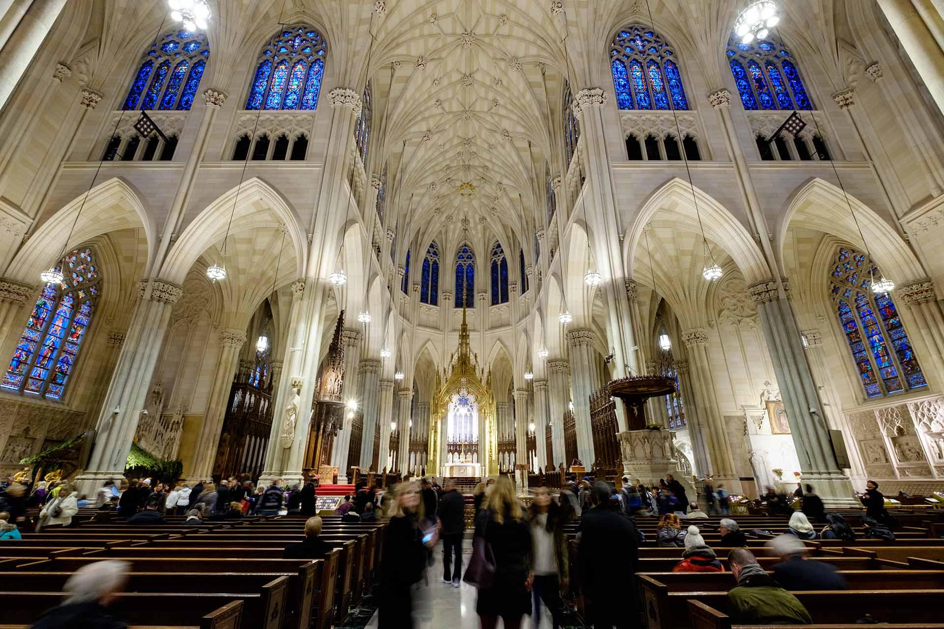 People walk and gather inside a grand cathedral with high vaulted ceilings, arched columns, and intricate stained glass windows. The pews are filled, and the altar is illuminated at the far end.