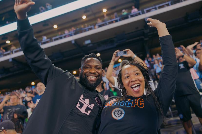 Two enthusiastic soccer fans smile and pose for the camera at a crowded stadium, one wearing a black Miami shirt and the other in a black NYCFC shirt, with cheering spectators in the background.