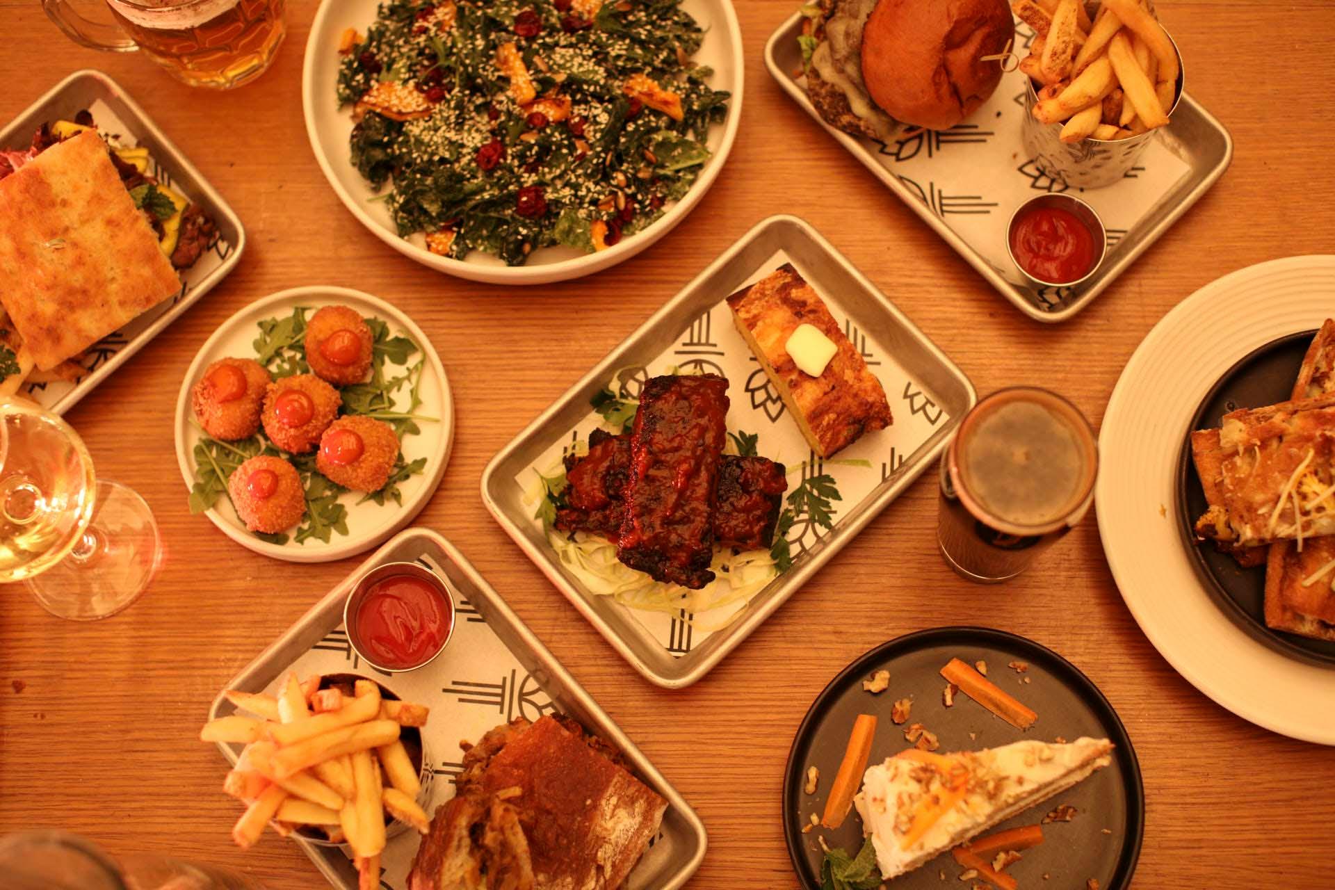 A variety of dishes on a wooden table, including fries, grilled meat, a salad, a burger, and dessert. Some plates have dipping sauces, and a drink is placed at the edge. The arrangement is casual and inviting.