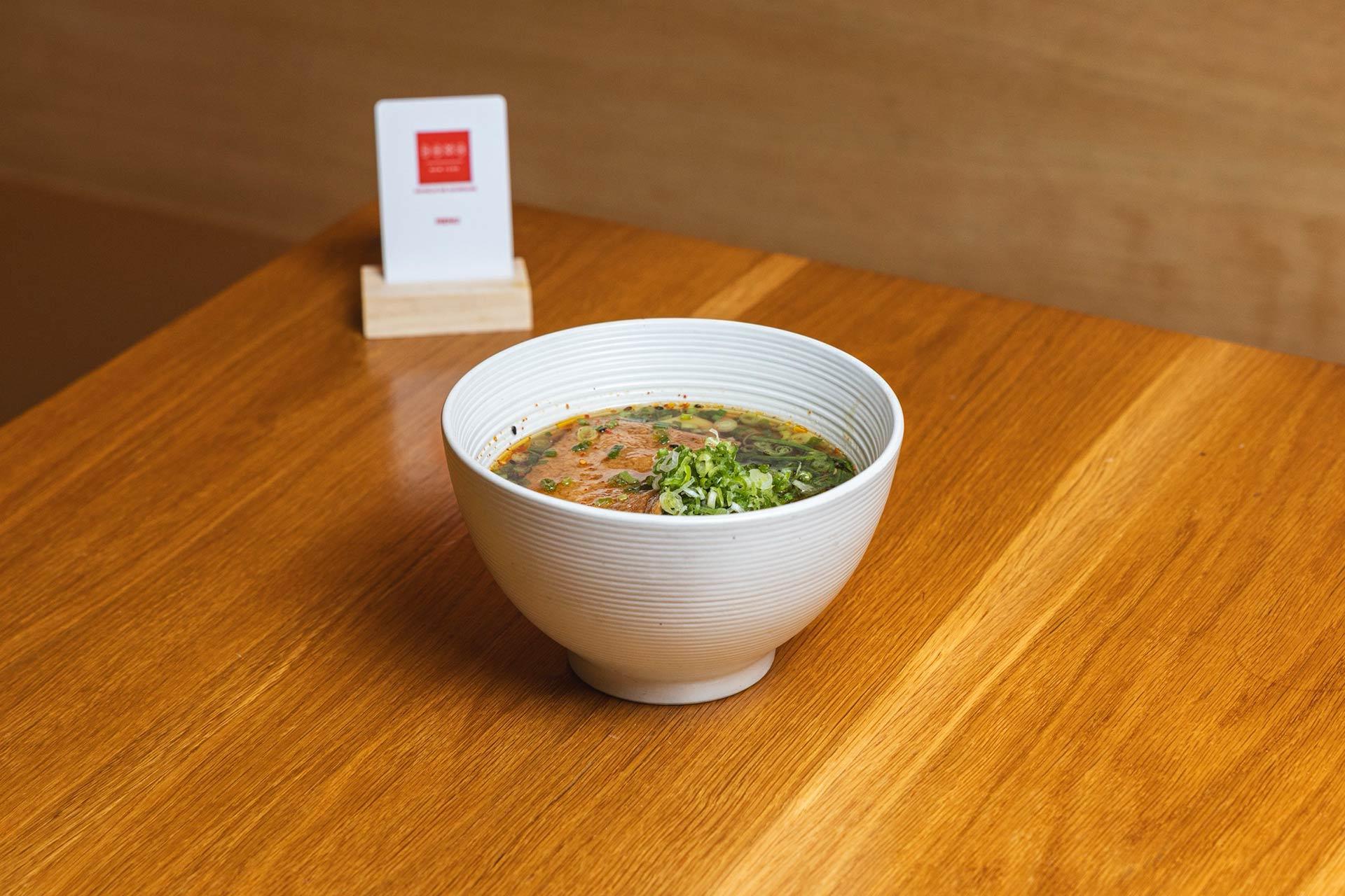 A white bowl of soup garnished with chopped green onions sits on a wooden table, with a small card holder in the background.