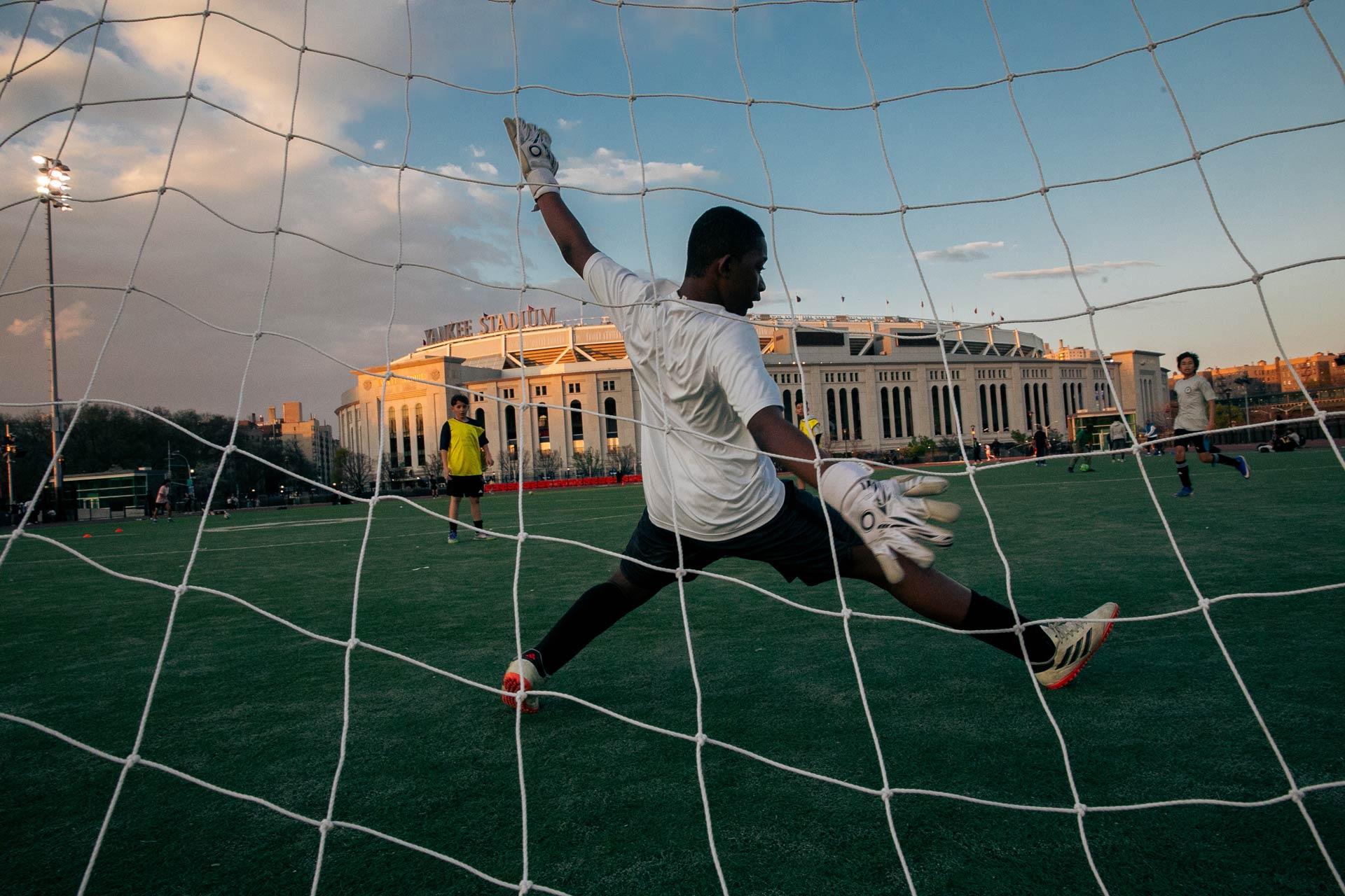 A soccer goalie, mid-air with arms outstretched, defends the goal as the sun sets behind a large stadium. Other players and the open field are visible in the background.