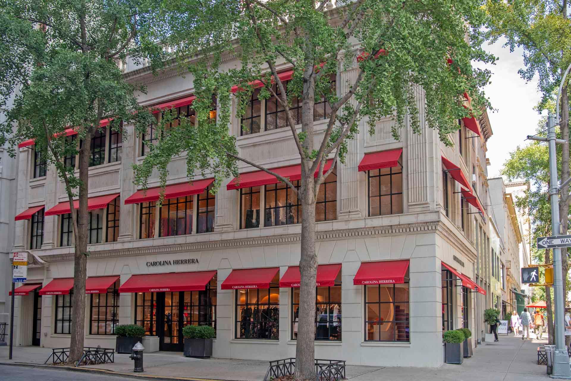 A corner building with cream-colored walls and red awnings, identified as a Carolina Herrera store. It features large windows and is surrounded by green trees on a sunny day. The street signs read "York Ave" and "East 69 St.