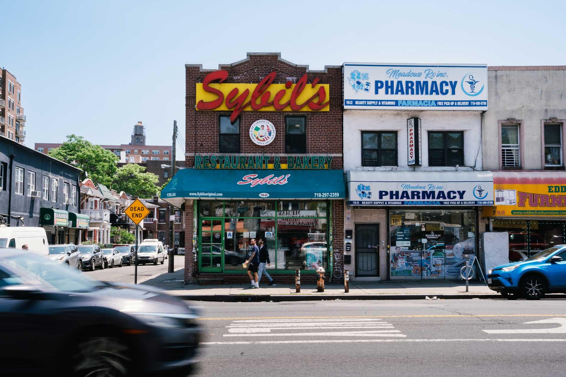A brick building with a large yellow and red “Sybil’s” sign houses a restaurant and bakery. Next door are two storefronts with “Pharmacy” signs. Cars and people are visible on the street in front.