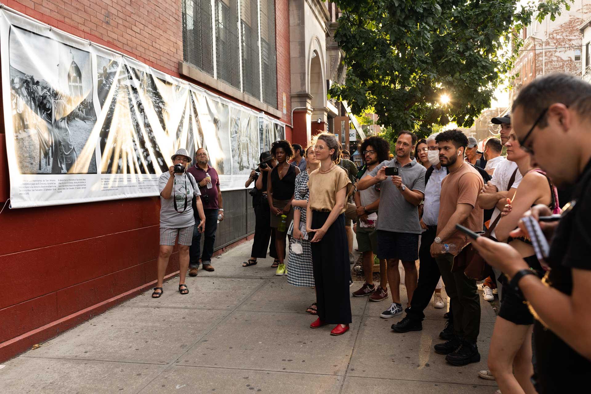 A diverse group of people stand on a city sidewalk, attentively viewing large black-and-white photographs displayed on a brick building’s exterior. Some hold cameras or phones; sunlight filters through nearby trees.