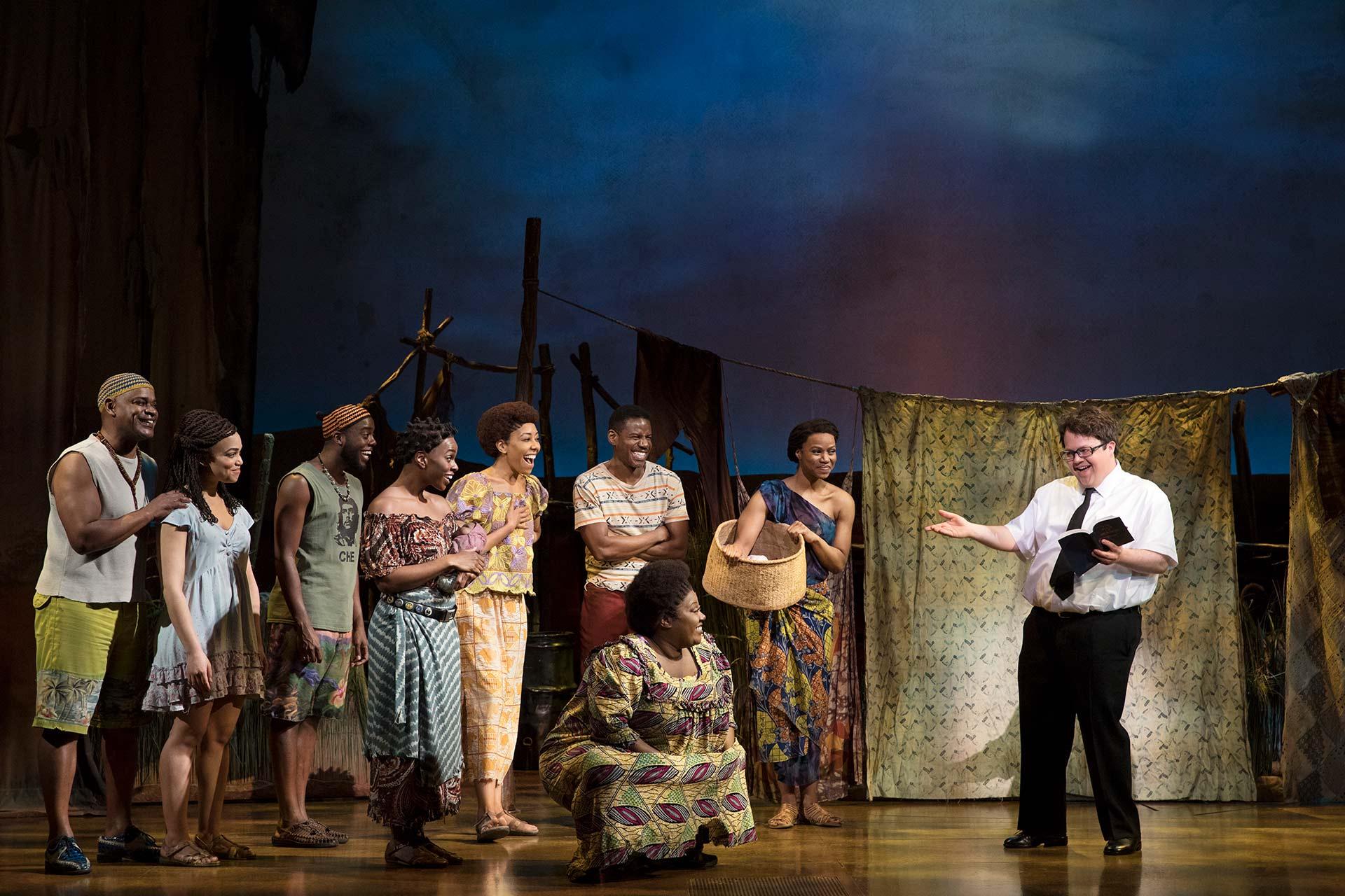 A group of people in colorful clothing stand on a stage, listening to a man in glasses holding a book and gesturing. The background suggests an outdoor village scene with draped fabric and props.