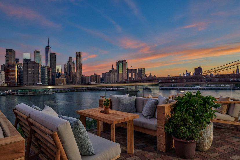 Rooftop patio with wooden furniture and potted plants overlooks a river and the New York City skyline at sunset, with the Brooklyn Bridge visible in the background.