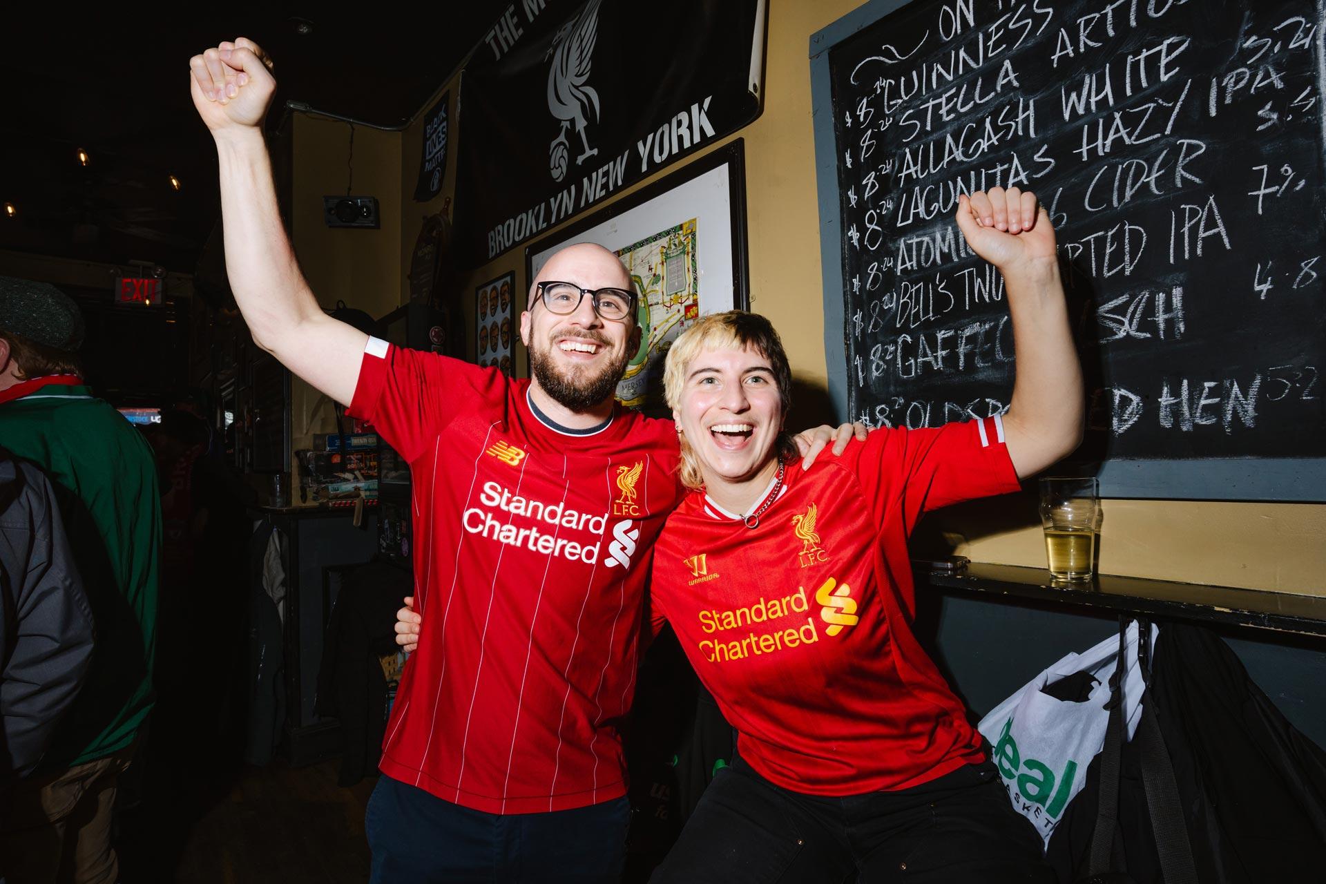 Two excited fans wearing red Liverpool jerseys celebrate with raised arms inside a bar, smiling at the camera. A chalkboard beer menu and drinks are visible in the background.