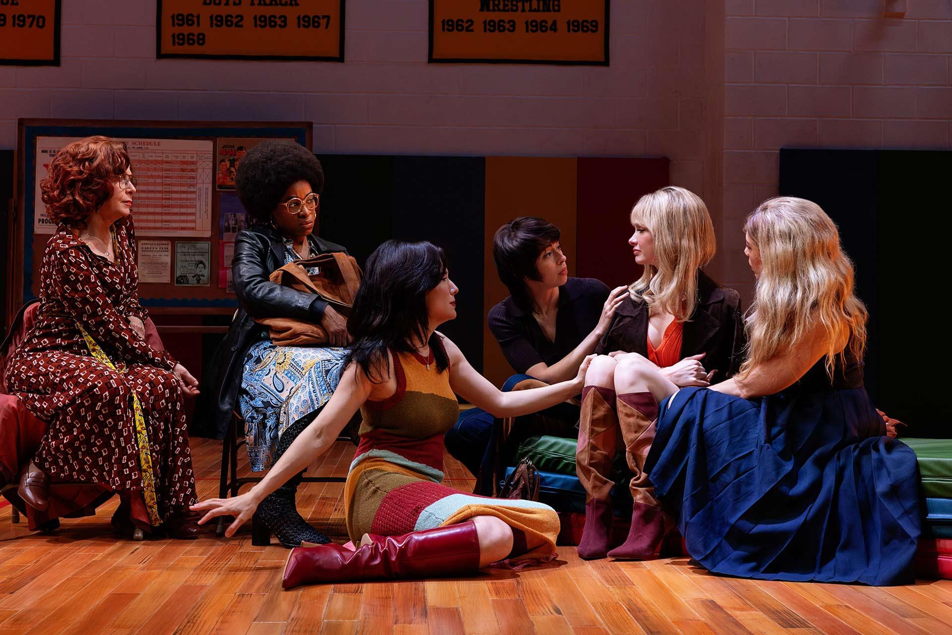 Six women sit in a circle on a gym floor, some reaching out or talking to each other. They wear colorful, retro-style clothing. Banners with years hang on the wall behind them. The scene appears dramatic and emotional.