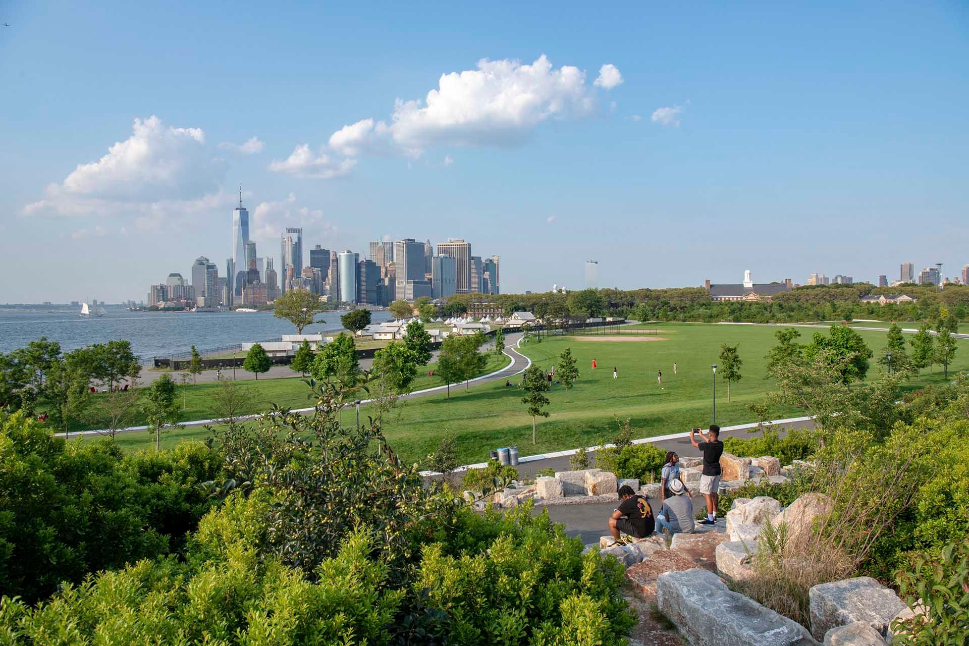People relax on rocks overlooking a large grassy park with paths, trees, and a city skyline featuring tall buildings in the background under a blue sky with scattered clouds.