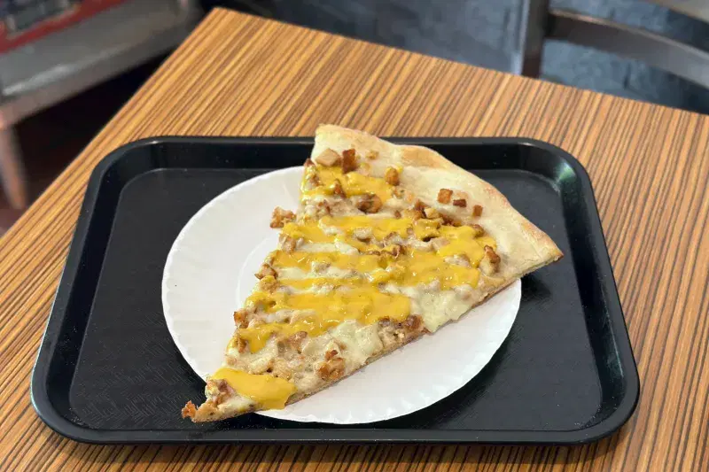 A single slice of pizza on a paper plate, sitting on a black plastic serving tray. The pizza has chunks of fried chicken cutlet as a topping, and a heavy drizzle of golden-yellow honey mustard sauce.