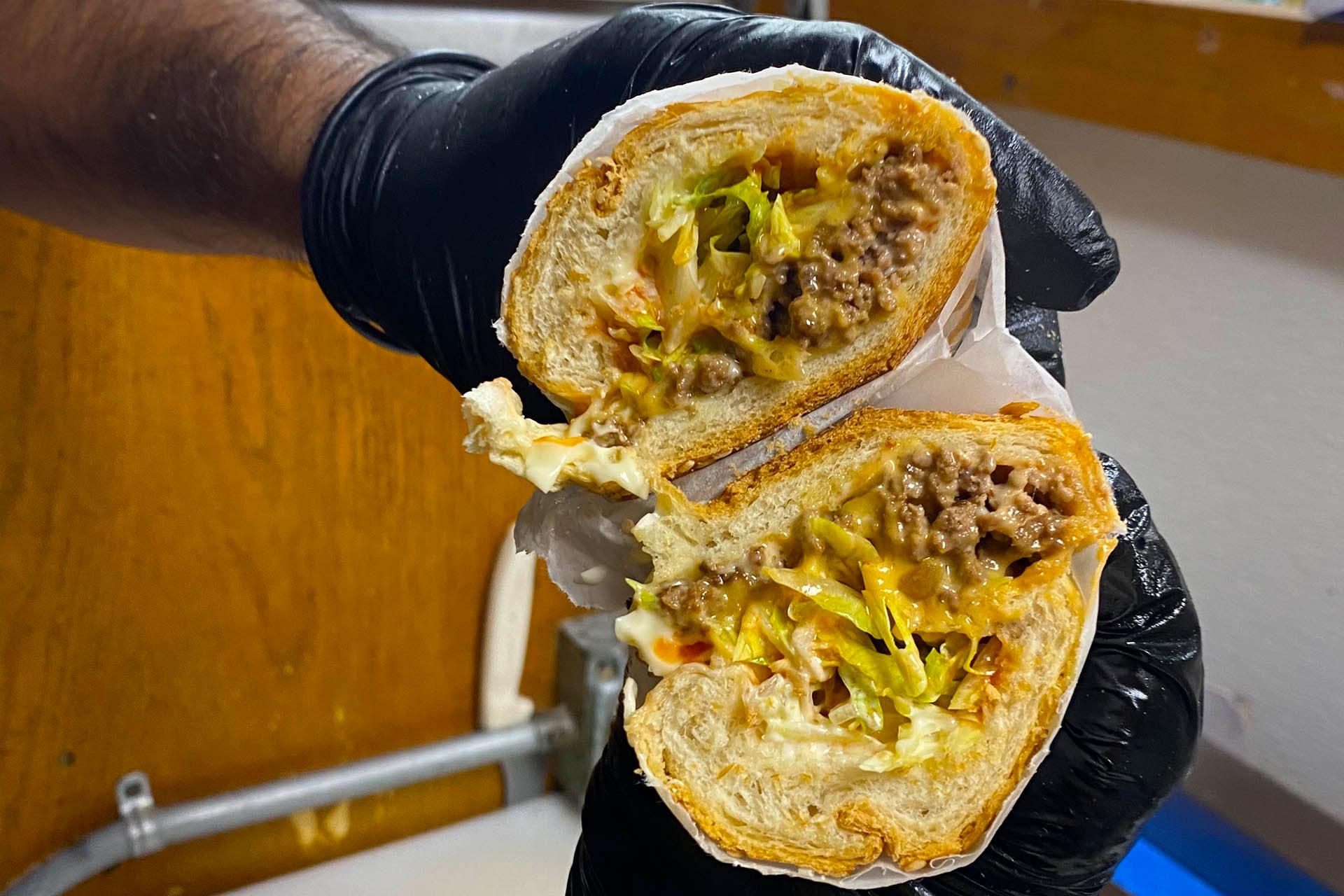 A close-up of a gloved hand holding a sandwich cut in half, filled with ground beef, shredded lettuce, melted cheese, and sauce on a toasted bun.