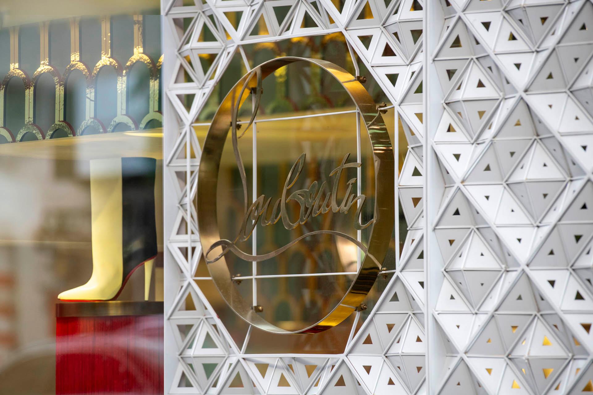 Storefront with a geometric patterned wall and a circular, gold logo sign featuring cursive text. Inside, a display showcases an elegant red high heel shoe.