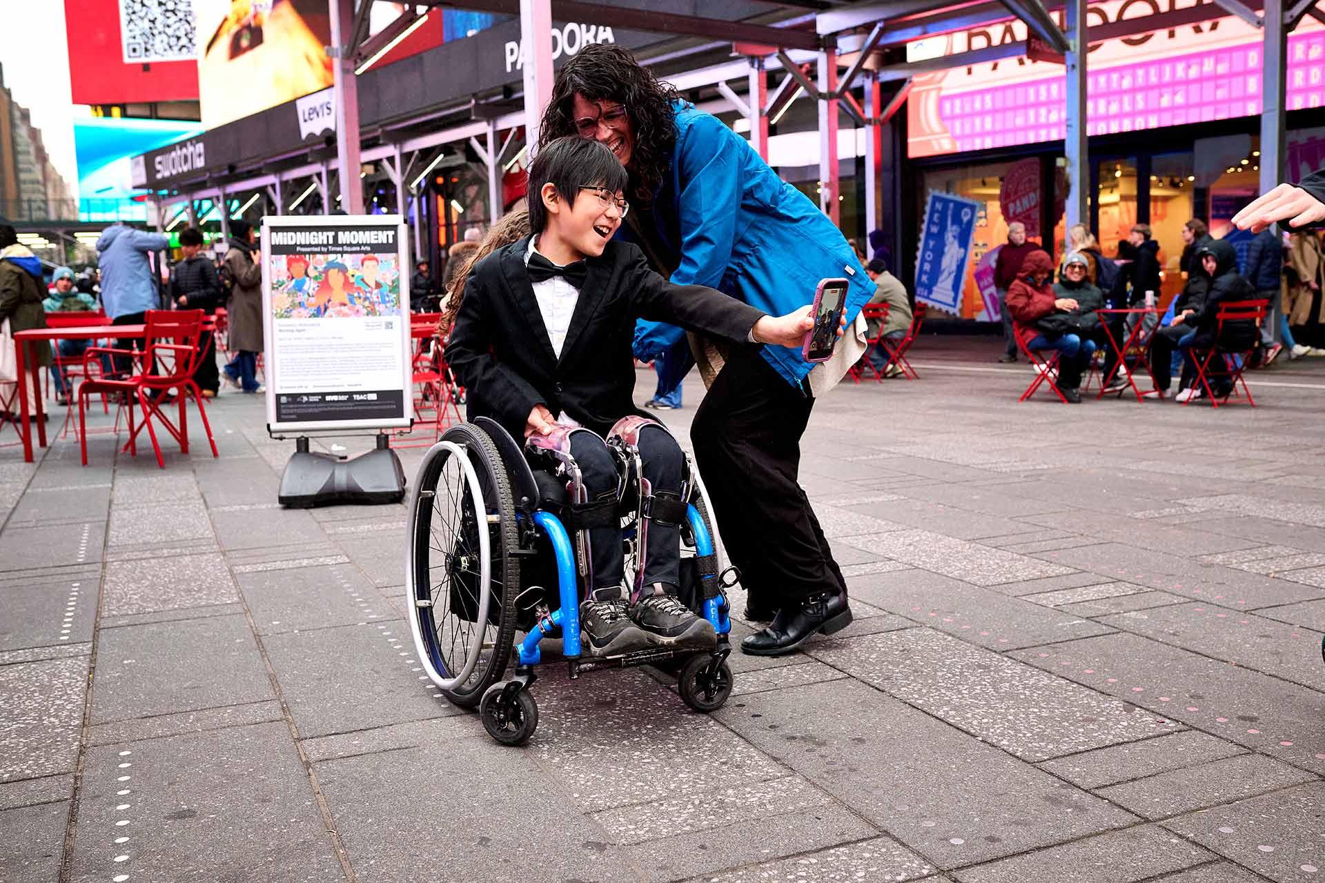 A smiling child in a wheelchair takes a selfie with a woman in a blue coat, surrounded by people sitting at red tables in a busy outdoor plaza with bright signs.