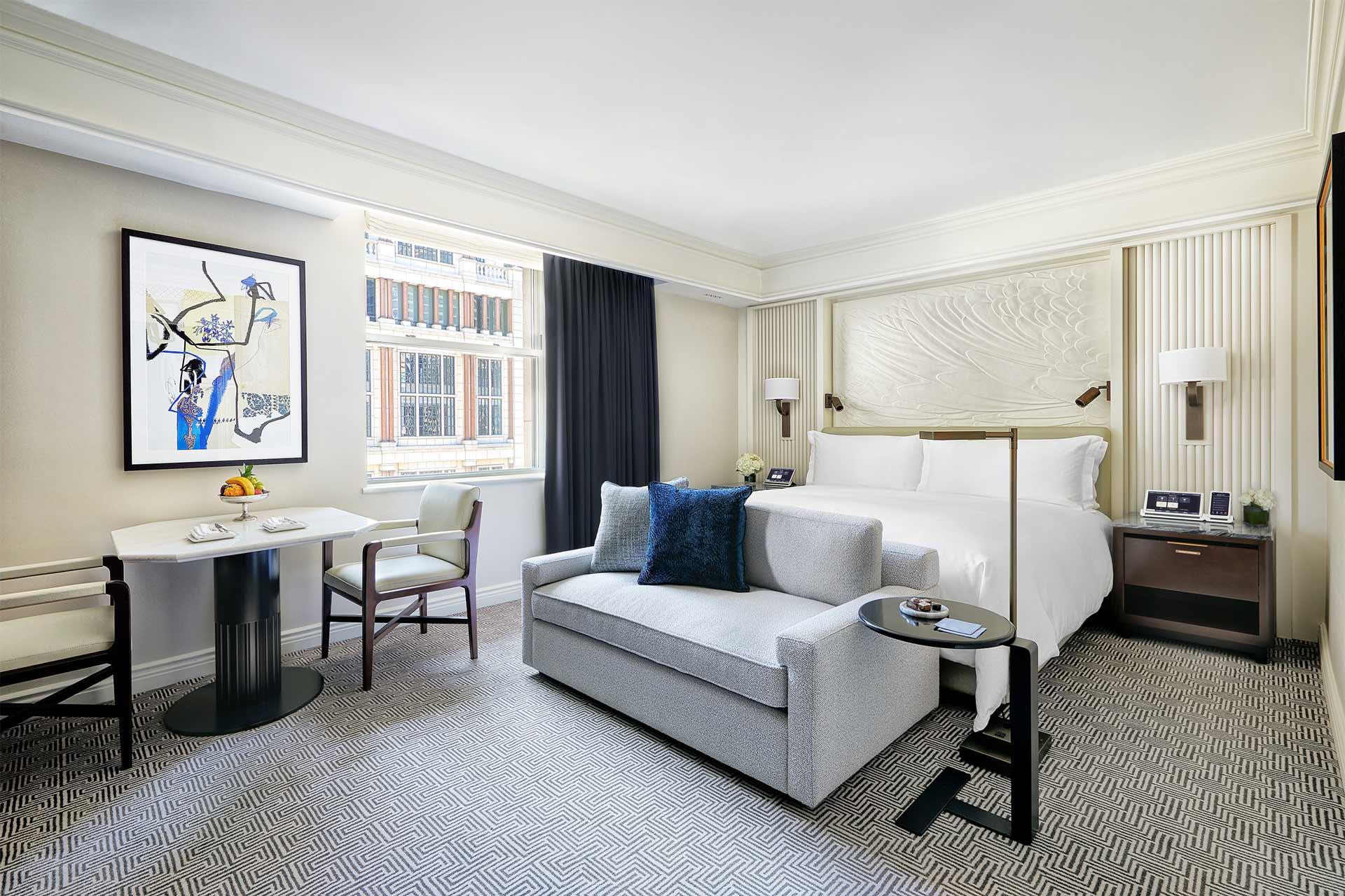 A hotel room with a large bed, light grey sofa, and a dining table with two chairs. A decorative painting hangs on the wall.