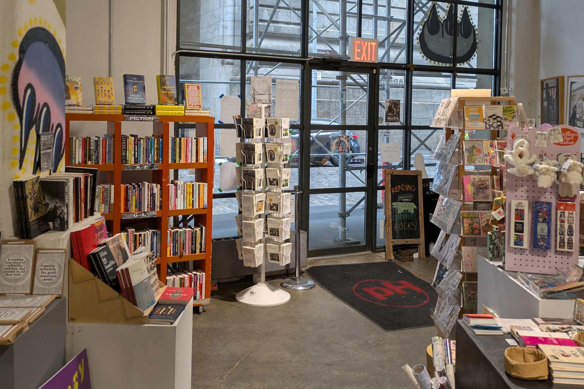 A cozy bookstore with shelves of books, greeting cards on a rotating stand, and assorted gifts. Large windows and a glass door show a city street and scaffolding outside. An exit sign hangs above the door.