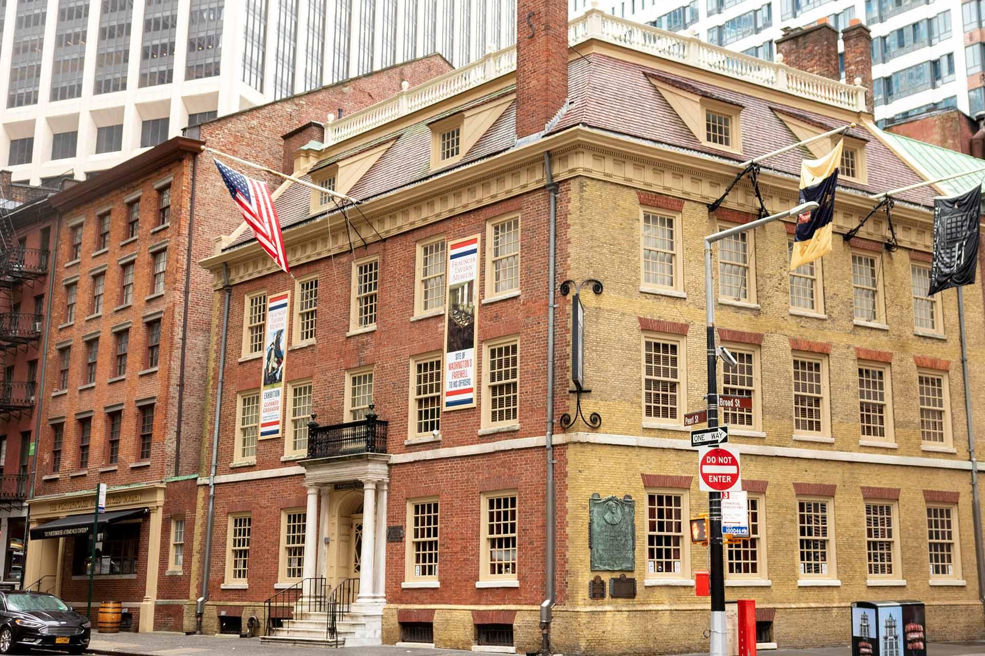 A historic brick building with cream trim and several flags displayed, located on a city street corner with modern skyscrapers in the background. A red "Do Not Enter" sign is visible at the intersection.