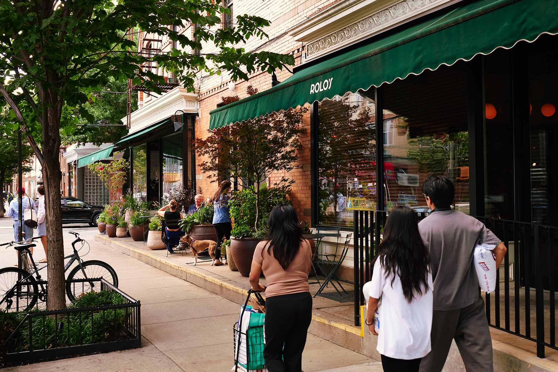 People walking on a sidewalk past Rolo’s restaurant, which has outdoor seating and a green awning.