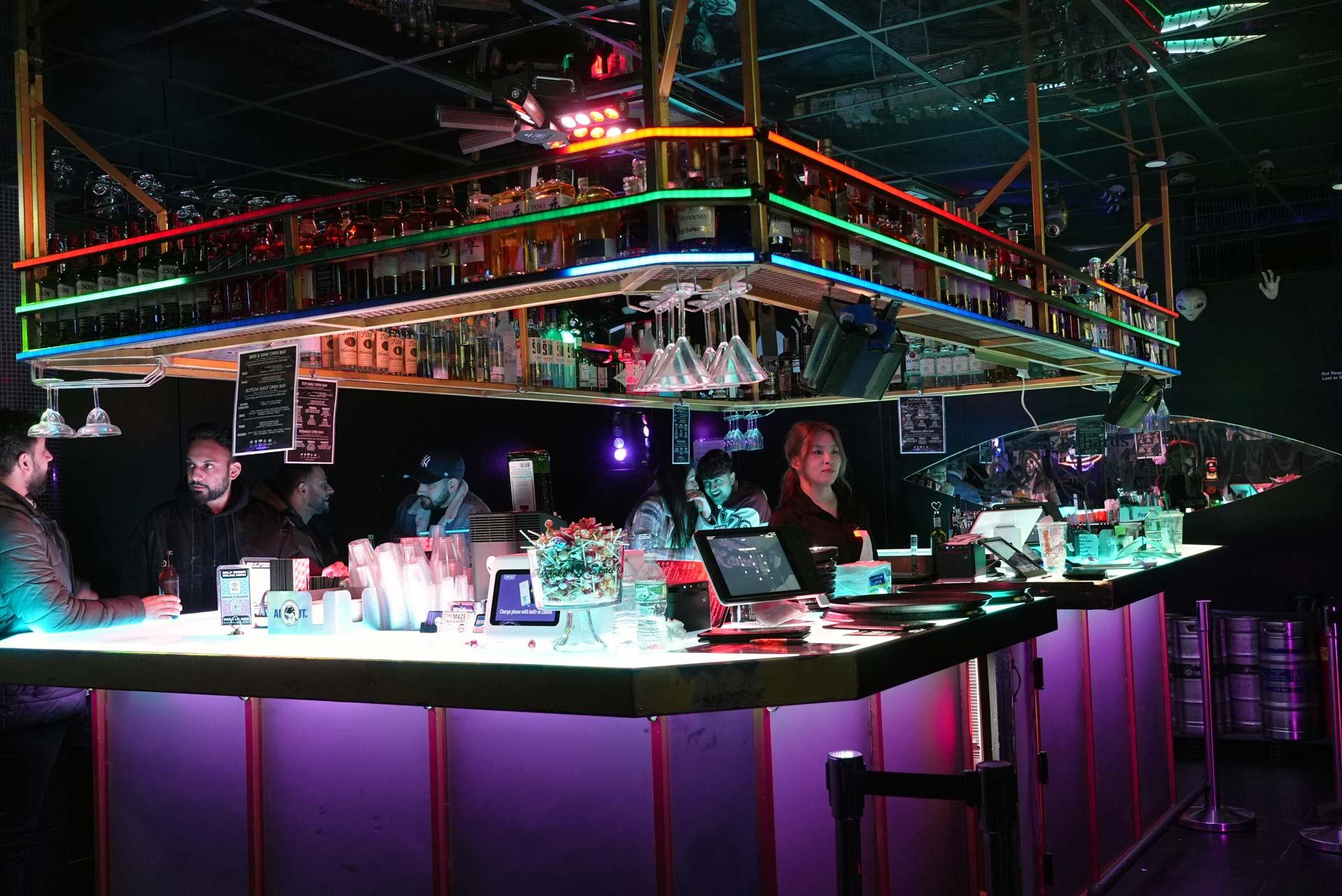 A neon-lit bar with shelves of bottles and hanging glasses. Several people stand or sit around the counter, while a bartender serves drinks. The atmosphere is lively with colorful lights and reflections.