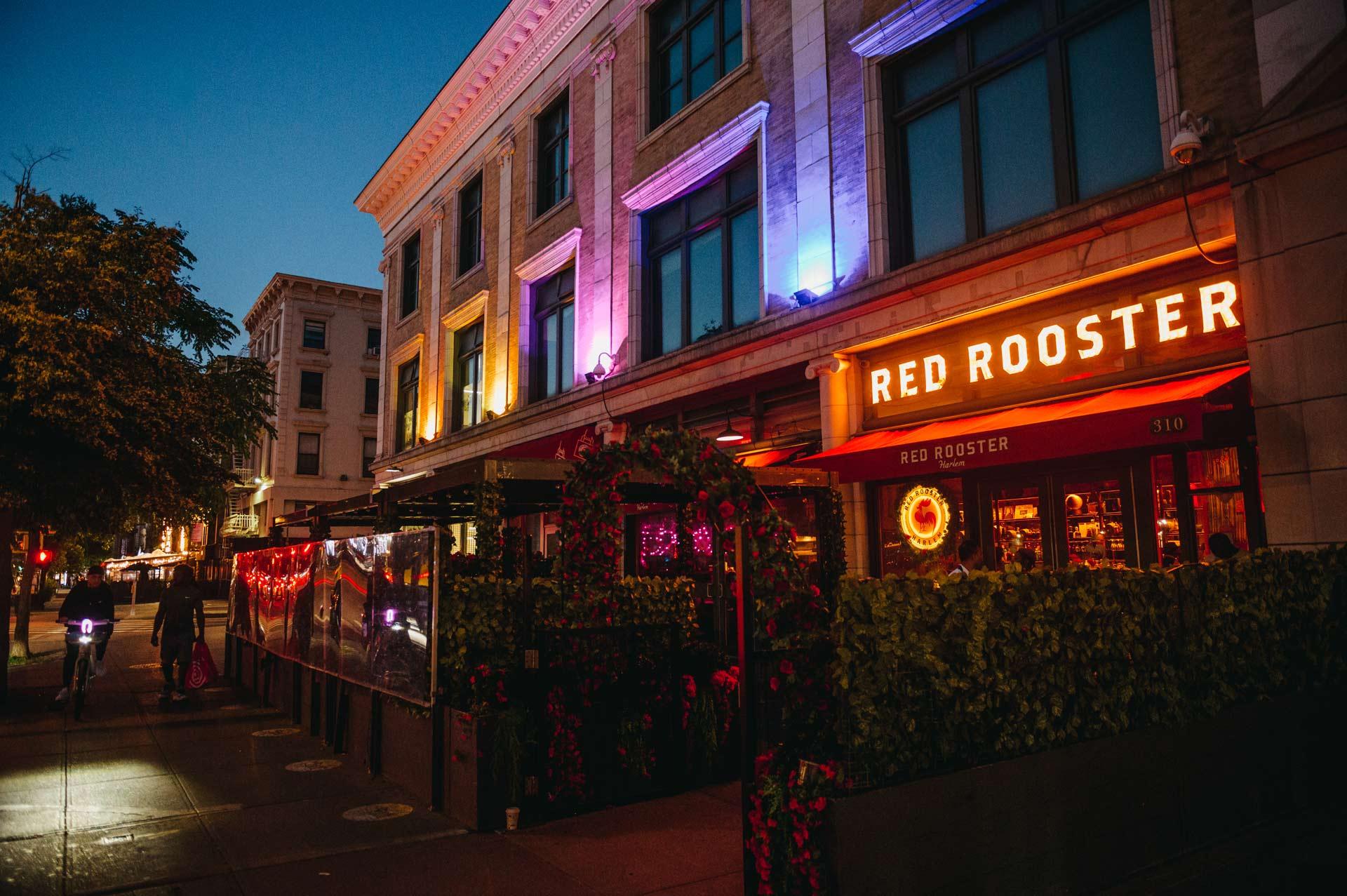 Street view of an illuminated restaurant at dusk named "Red Rooster." The building has a warm glow with colored lights and outdoor greenery.
