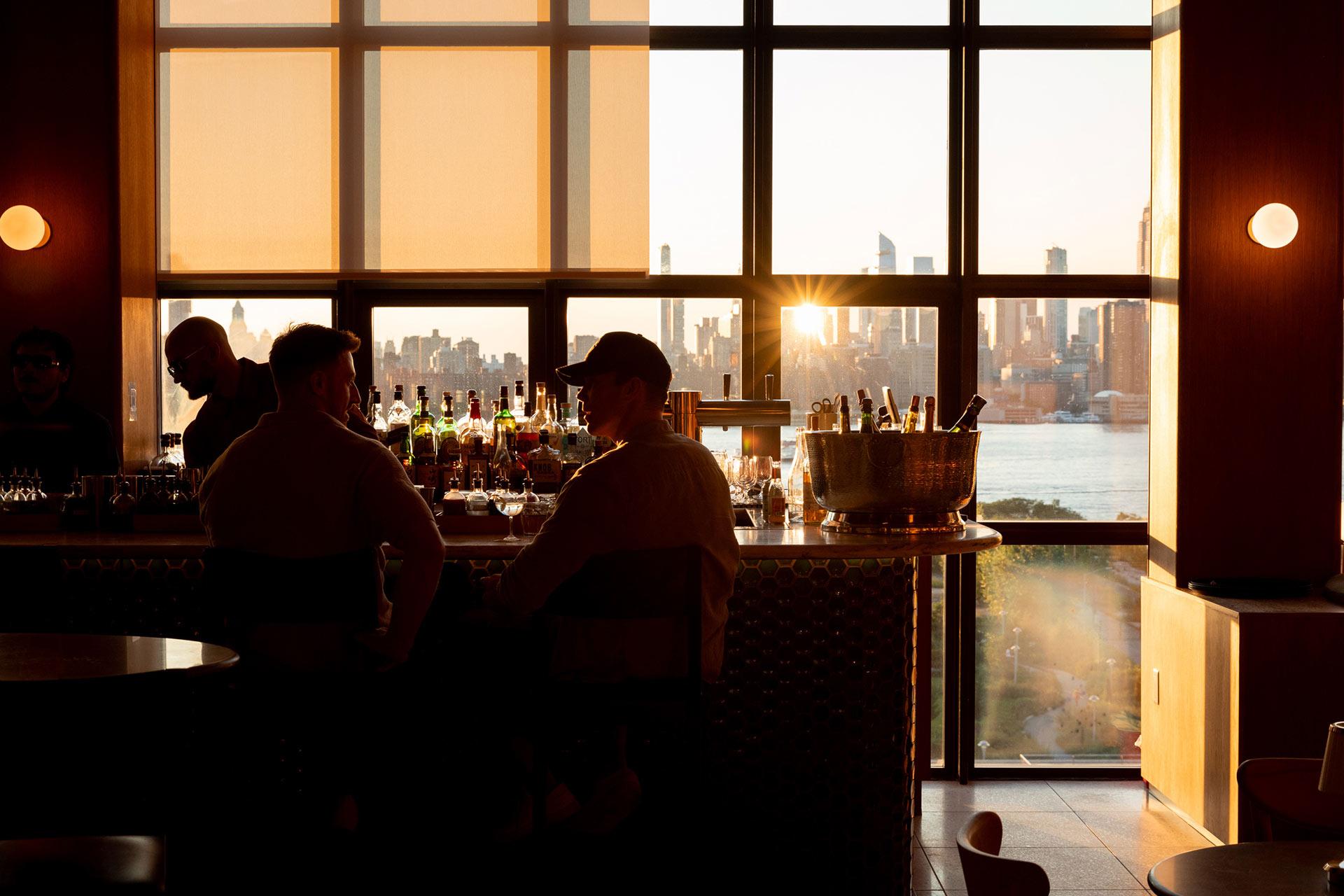 Two people sit at a bar in a dimly lit room with large windows; sunlight streams in, revealing a city skyline and river outside. Bottles and glasses are visible on the bar counter.
