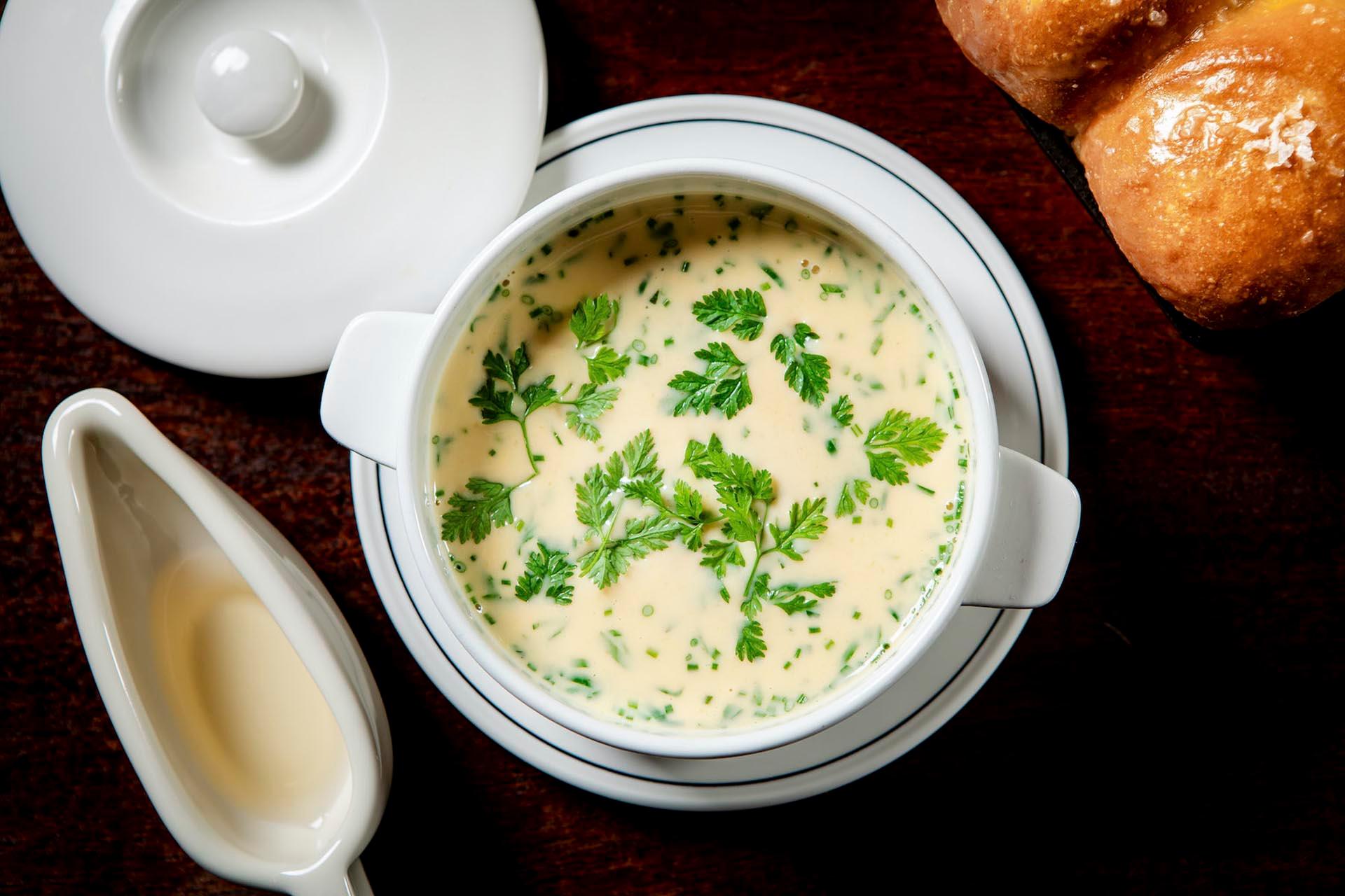 A bowl of creamy soup garnished with fresh herbs, served in a white dish. A small white sauce pitcher is nearby, and freshly baked bread is partially visible on a dark wooden surface.