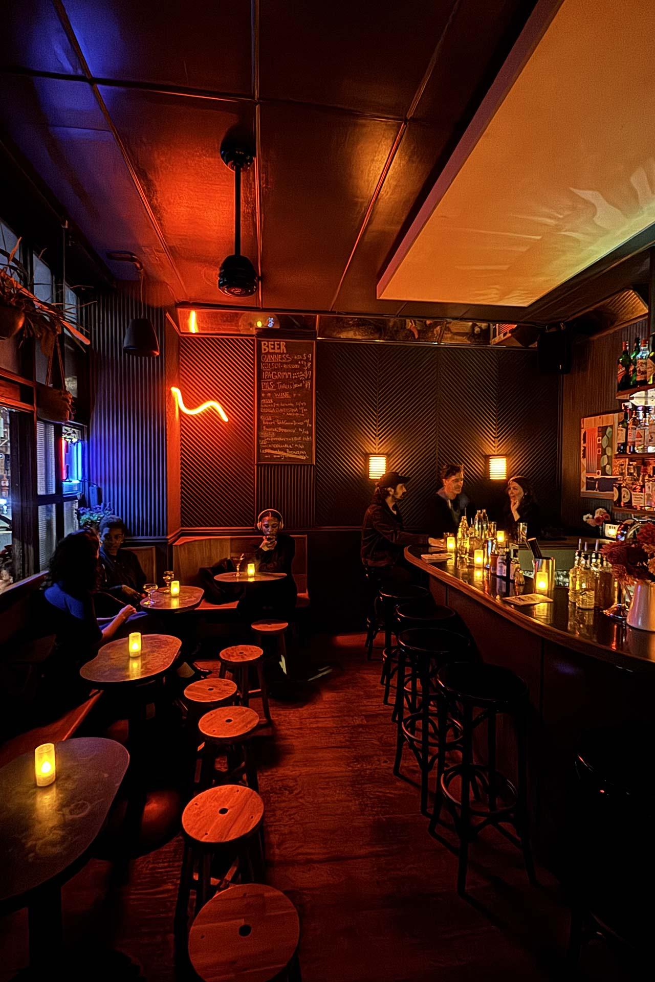Interior view of a dimly lit bar with patrons seated at small tables and the service counter, featuring a menu board on the wall and warm ambient lighting.