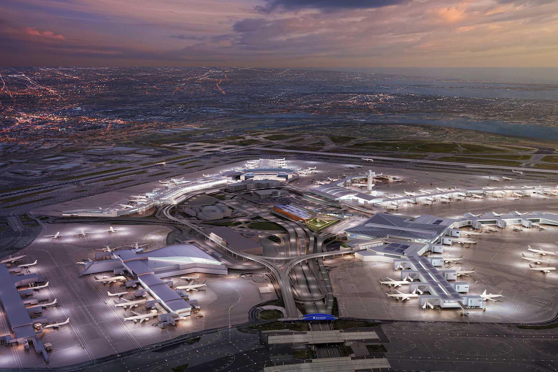 Aerial view of a large, brightly lit airport at dusk, with multiple terminals, airplanes parked at gates, runways, and a cityscape in the background under a cloudy sky.
