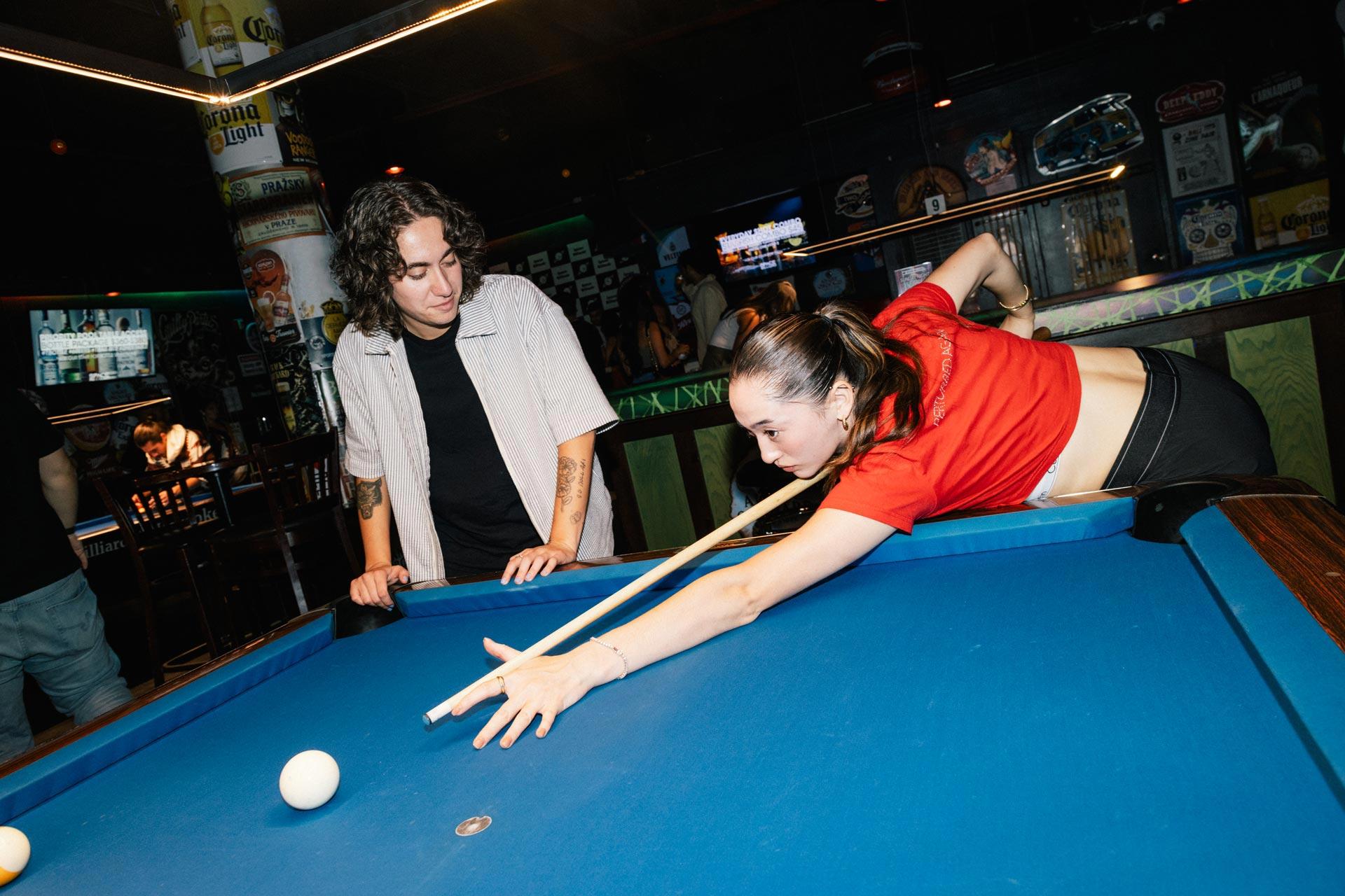 A young person in a red shirt lines up a shot at a blue pool table while another person in a light shirt watches them, both inside a dimly lit bar with posters on the walls.