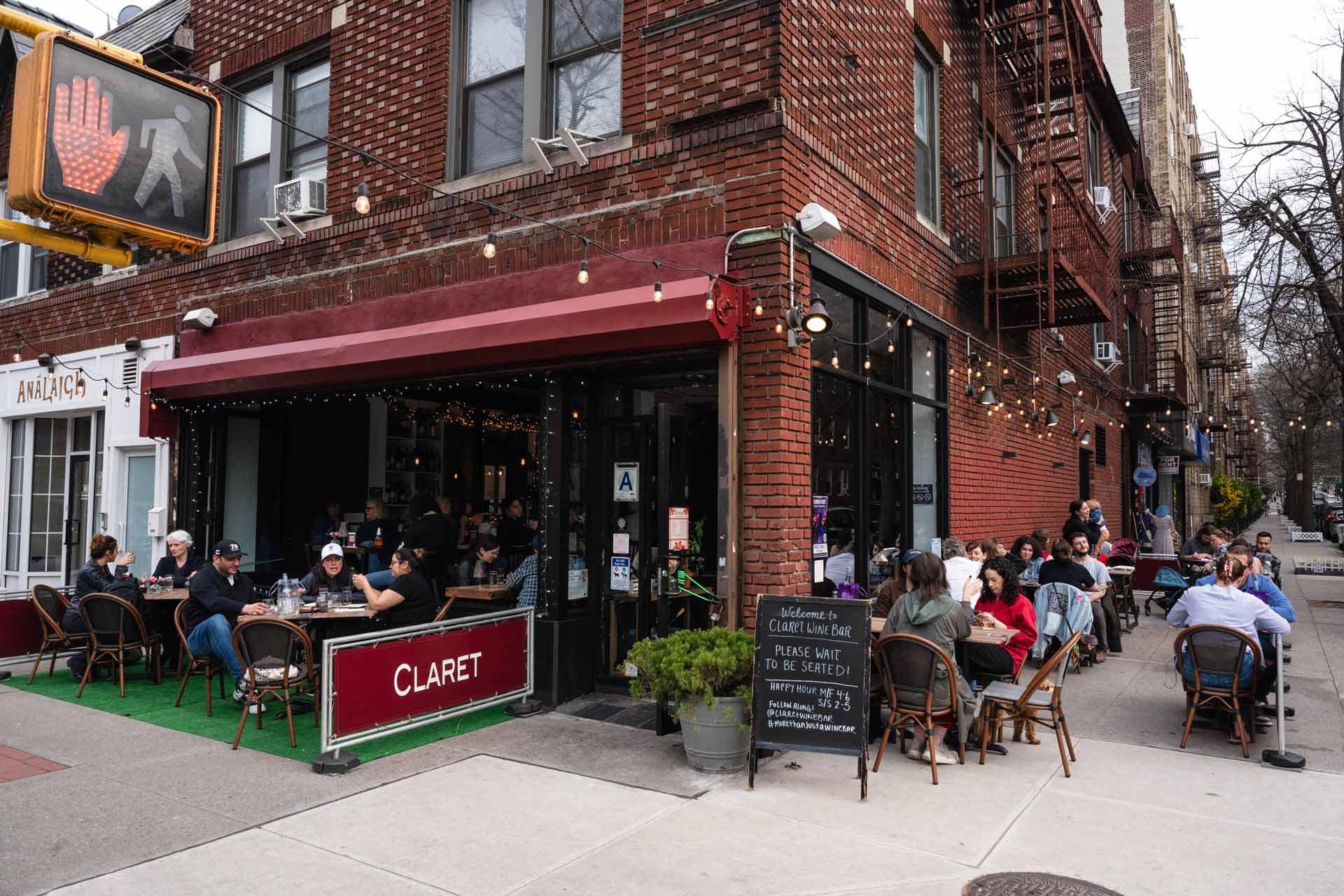People dine outdoors at a sidewalk café called Claret on a city corner. The café has string lights, a red awning, and a chalkboard menu. A traffic signal and brick apartment buildings are visible in the background.