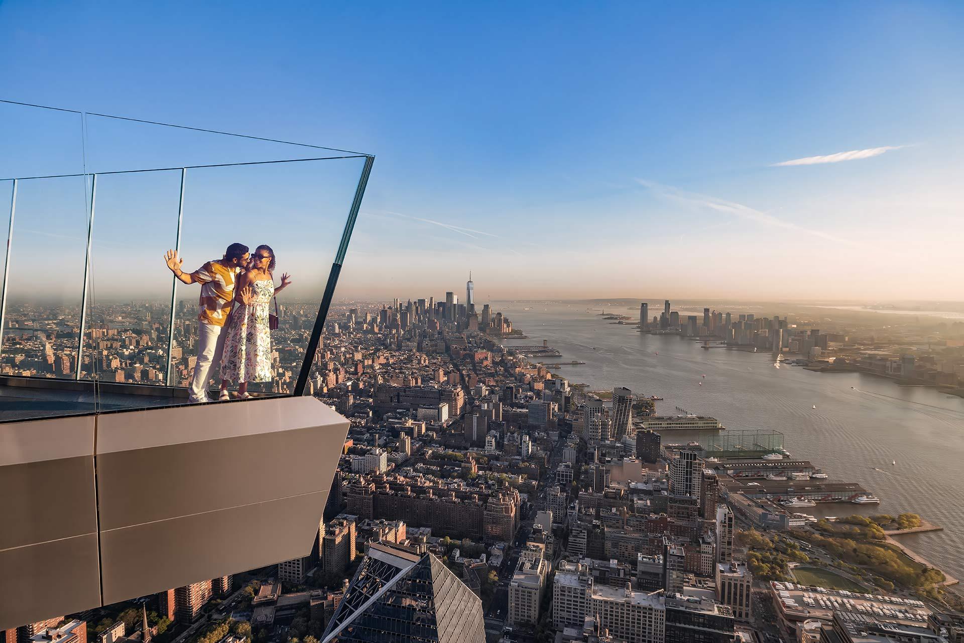 A couple stands on a glass observation deck high above New York City, overlooking skyscrapers, the Hudson River, and the distant skyline under a clear blue sky.