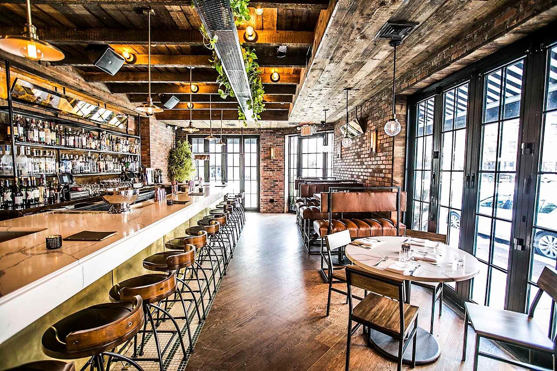 A rustic bar interior with a long marble counter lined with stools, exposed brick walls, and wooden ceiling beams. The space features leather booth seating on the right and large windows letting in natural light.