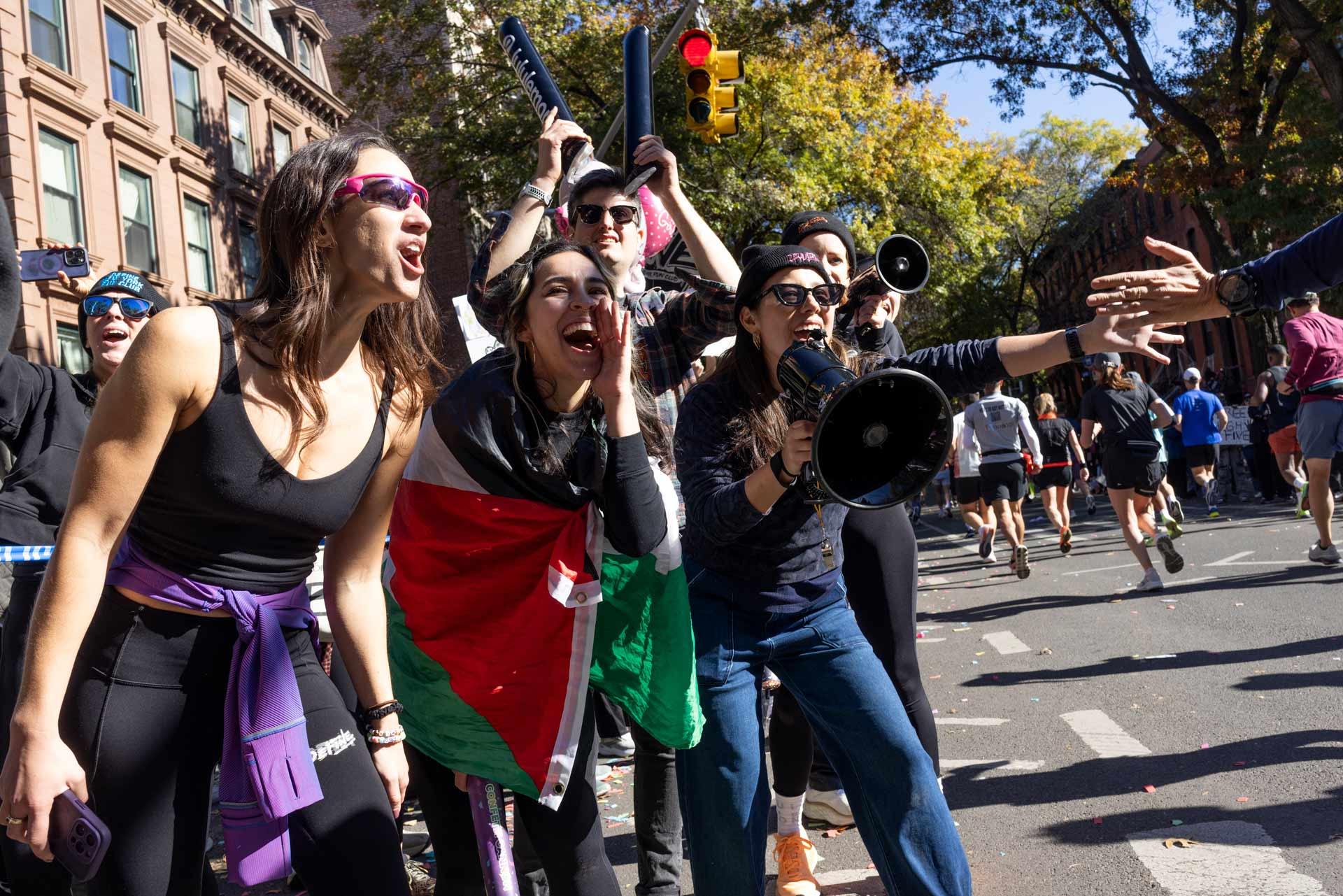 A group of enthusiastic spectators cheer at a marathon. One person waves a flag, and another holds a megaphone. There are autumn trees and buildings in the background.