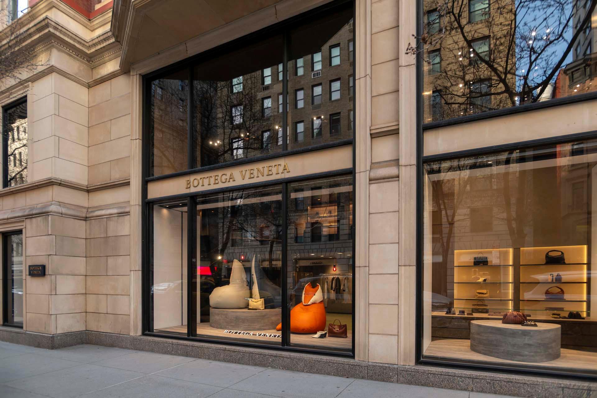 Street view of a Bottega Veneta store, displaying large windows with handbags and accessories. The interior showcases minimalist decor with a focus on luxury items. Reflections of nearby buildings and bare trees are visible in the glass.