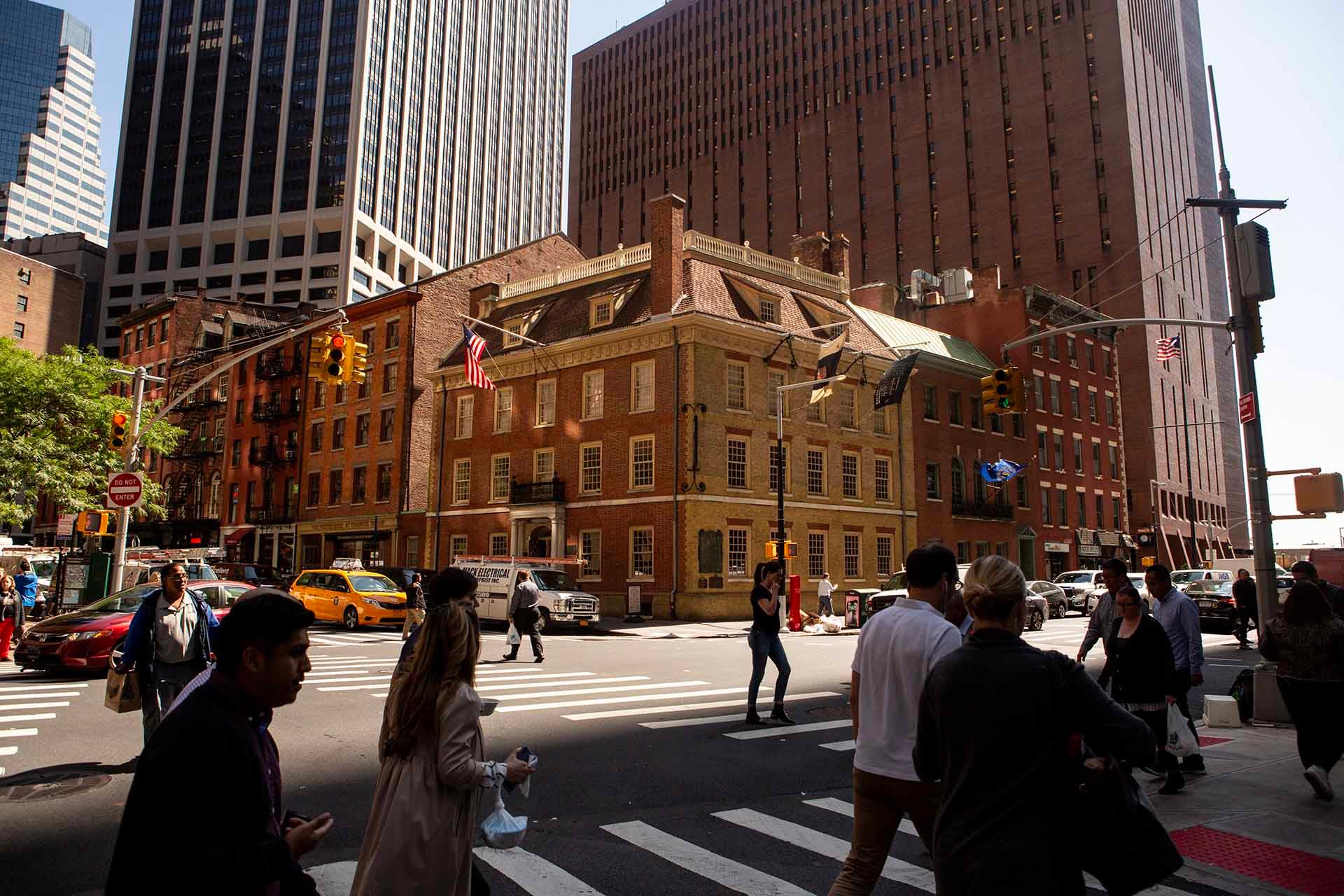 People cross a busy city street past Fraunces Tavern, a historic brick building with American flags, surrounded by tall modern office buildings in New York City on a sunny day.