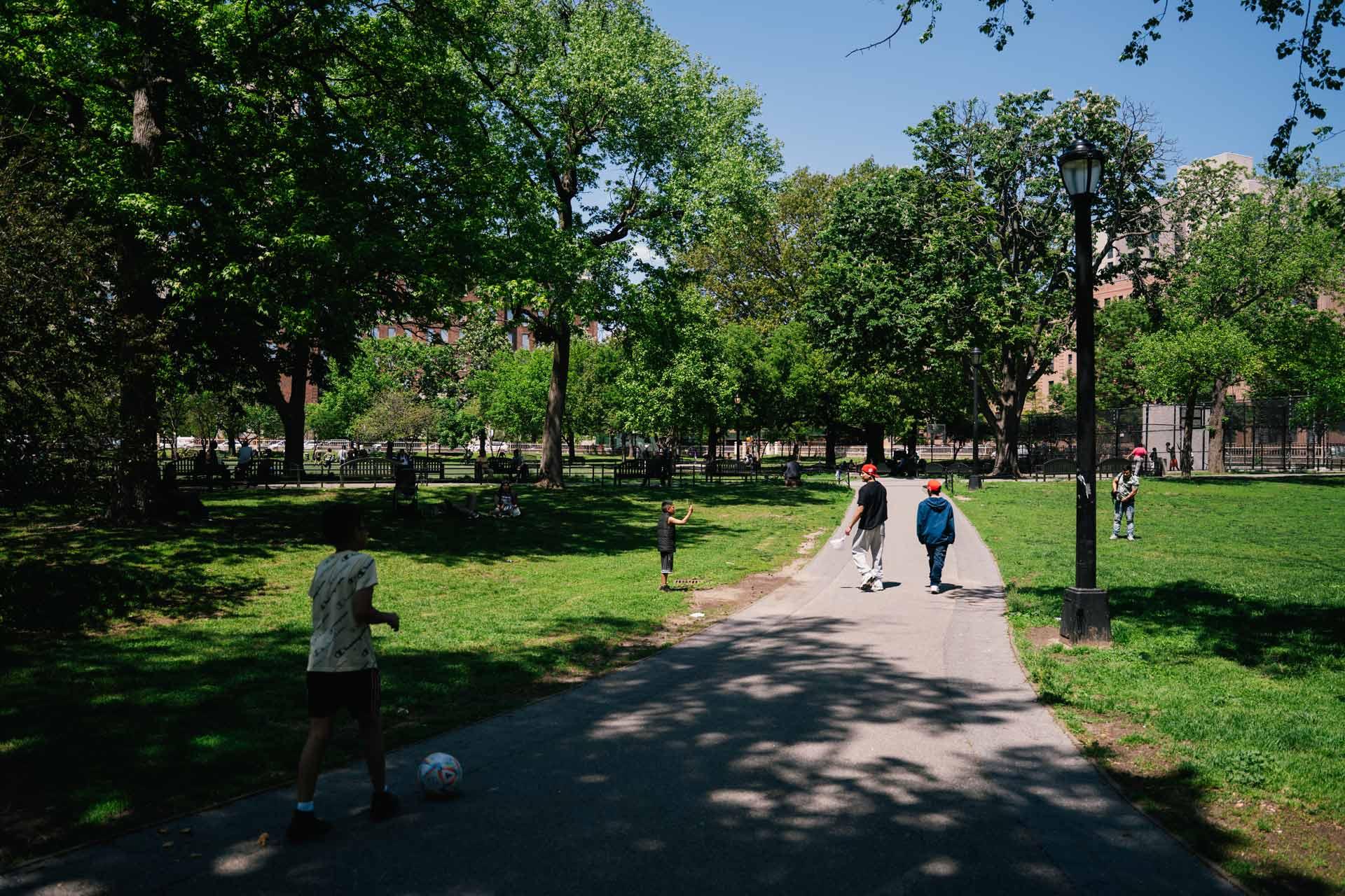 People walk and play in a green, sunlit park. Trees provide shade, and a few kids with a soccer ball stand on a paved path. A lamppost and fence are visible, with more people in the distance.