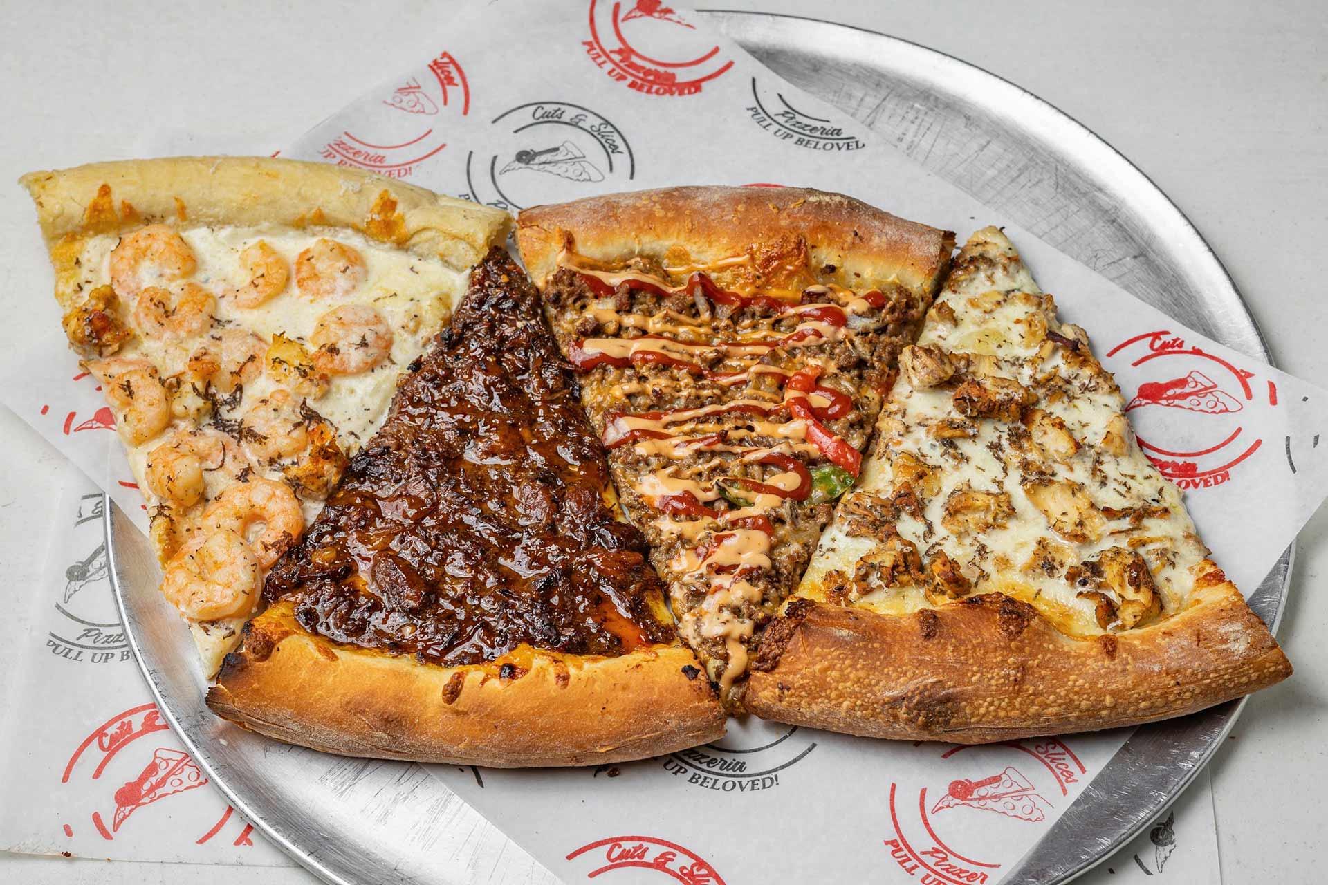Four assorted pizza slices with different toppings are arranged on a round metal tray lined with branded paper. The toppings include shrimp, barbecue sauce, ground meat, and chicken with herbs.