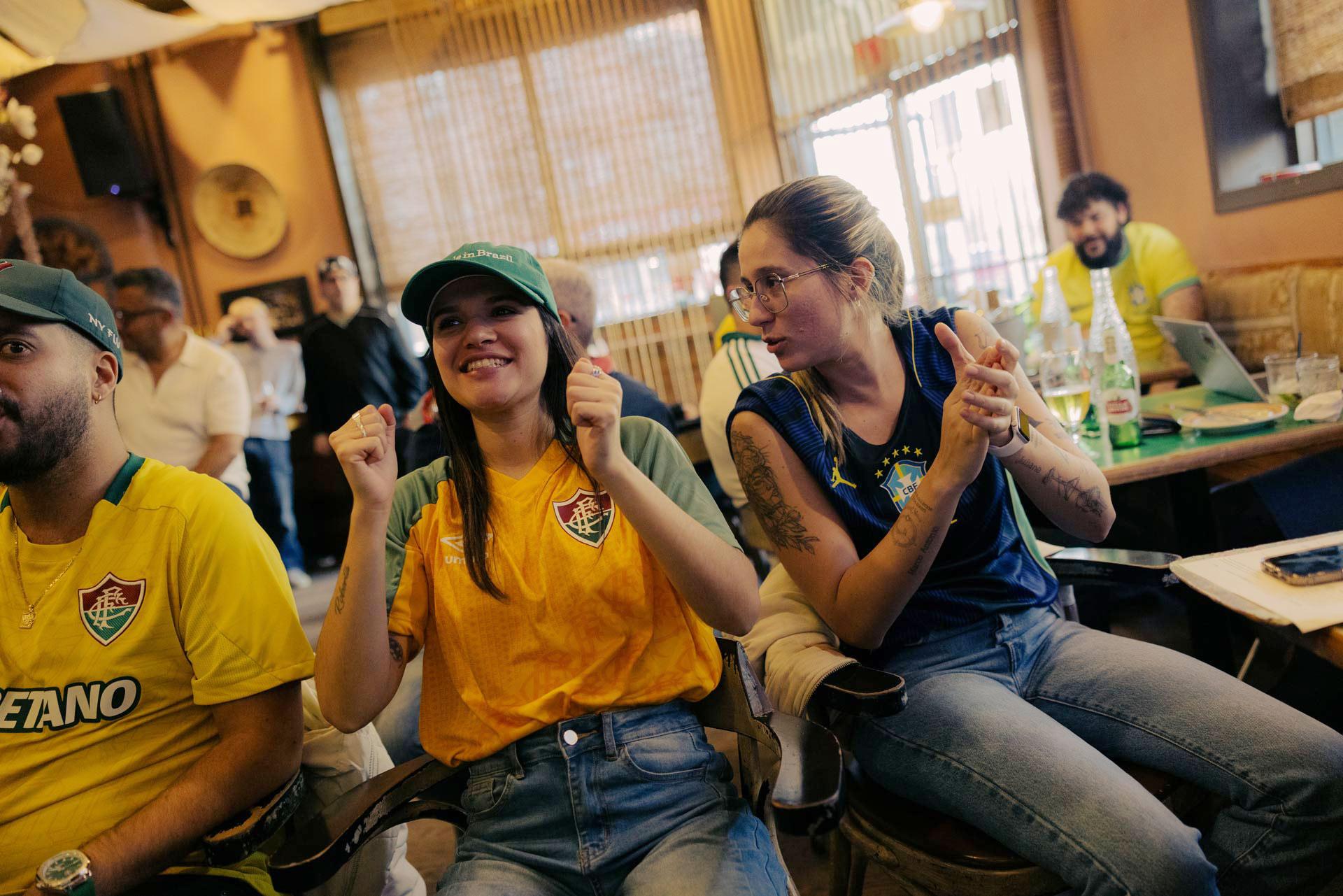 Two people sit indoors, wearing yellow and blue football shirts, cheering and clapping as they watch a Brazilian soccer game. Other people in football attire are visible in the background.