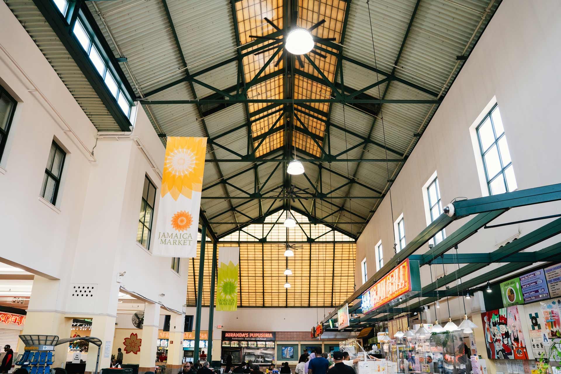 Indoor view of Jamaica Market with a high, triangular ceiling, hanging banners, food stalls on the right, and people walking around. Bright natural light comes through large windows at the end.
