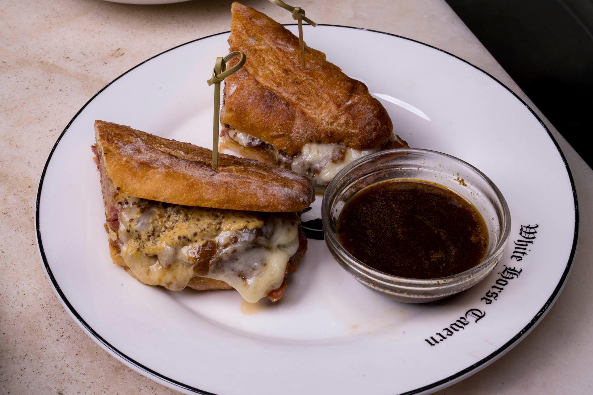 A plate with two halves of a sandwich filled with melted cheese and meat, secured with toothpicks. A small glass bowl of dipping sauce is beside the sandwich, all served on a white plate with "White House Tavern" text.