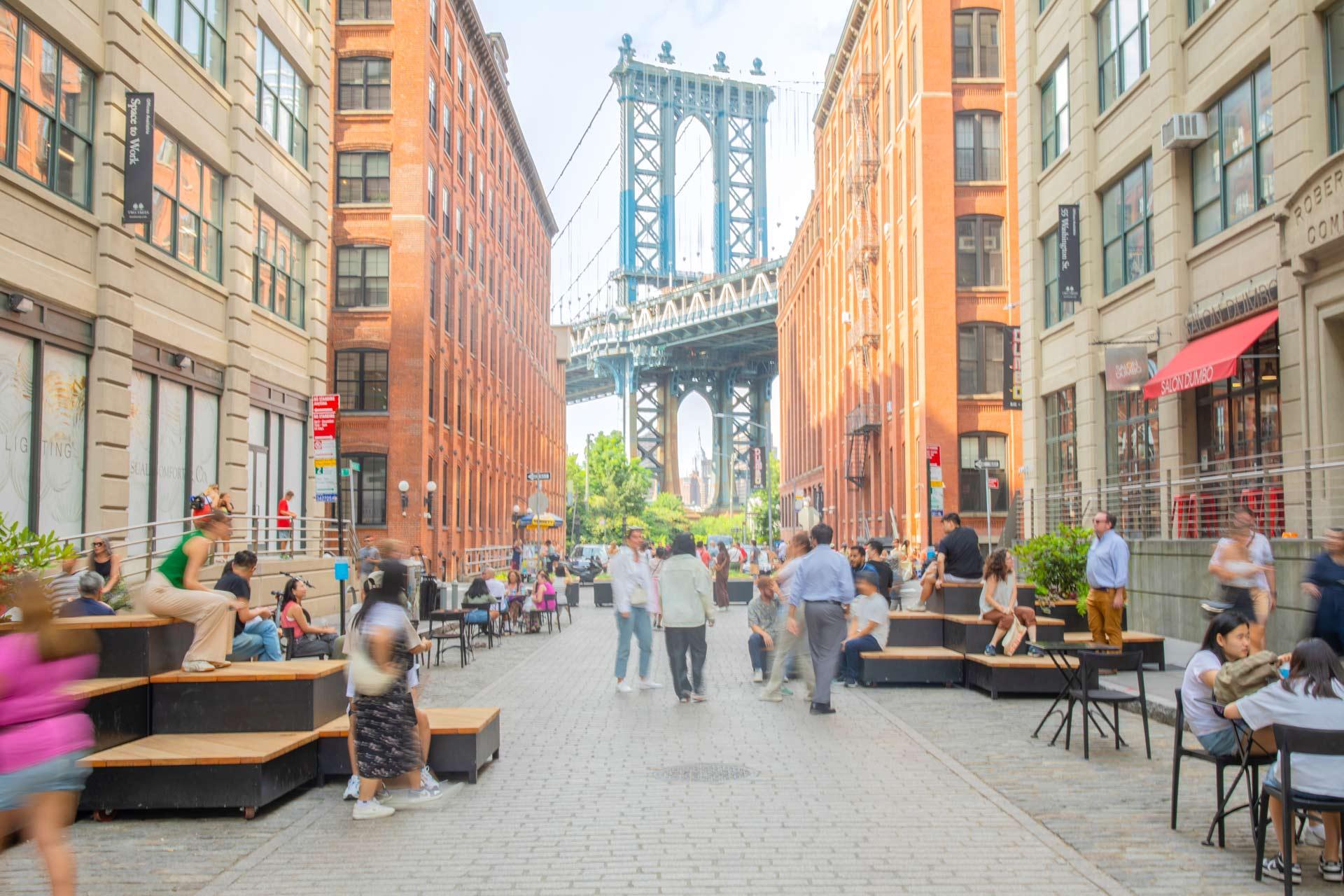 A busy cobblestone street in Brooklyn, New York, with people sitting and walking, tall brick buildings on both sides, and the Manhattan Bridge visible in the background.