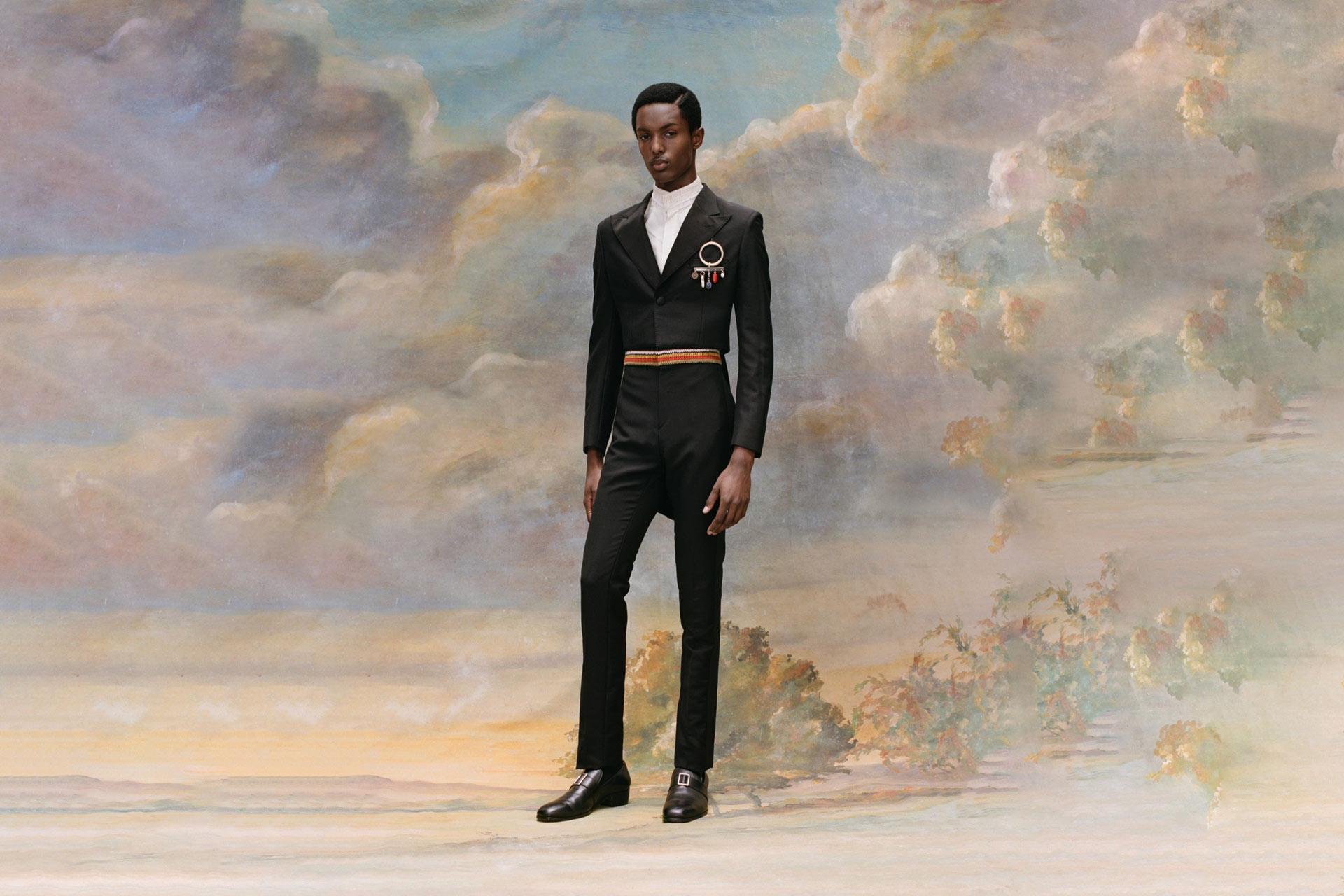 A person in a tailored black suit stands in front of a scenic painted background with clouds and foliage. They wear a high-collared white shirt, colorful belt, and brooch, exuding elegance and style.