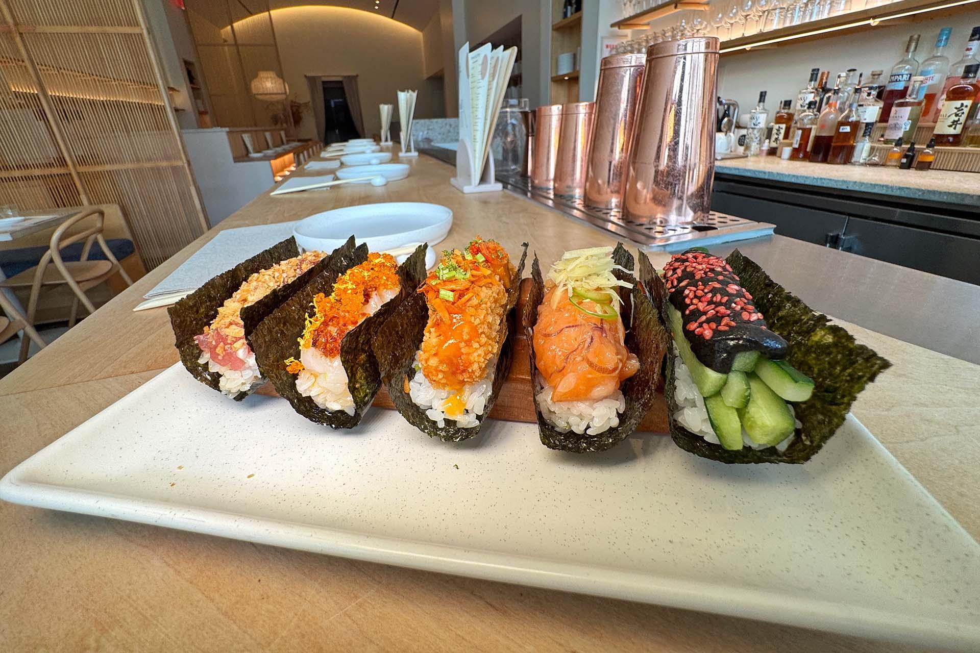 A row of six hand-rolled sushi temaki with various colorful fillings, arranged on a rectangular white plate atop a wooden counter in a modern, minimalist restaurant setting.
