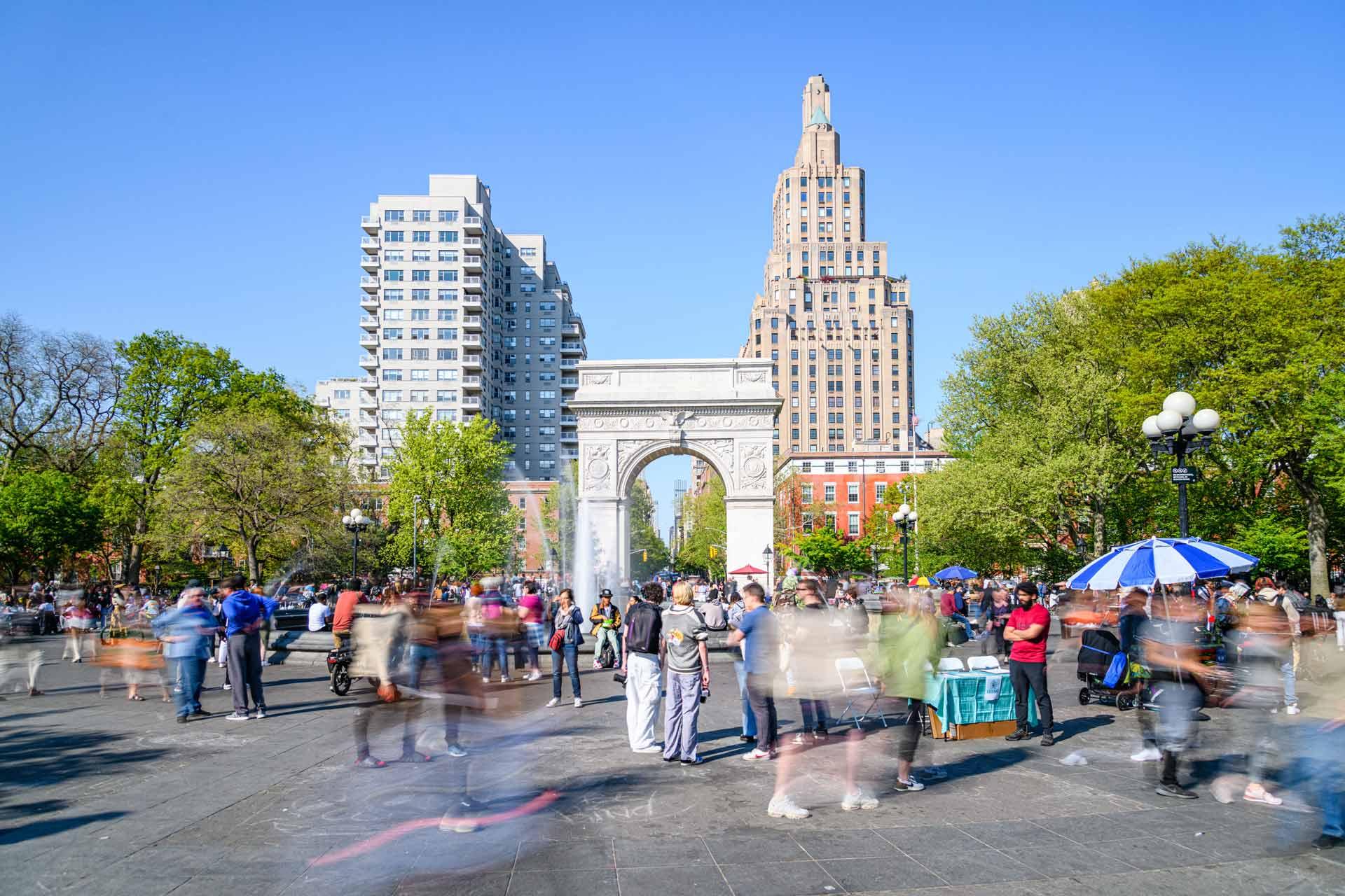 A bustling scene at Washington Square Park on a sunny day. People are walking and gathering near the iconic arch. Surrounding the park are tall buildings and lush green trees under a clear blue sky.