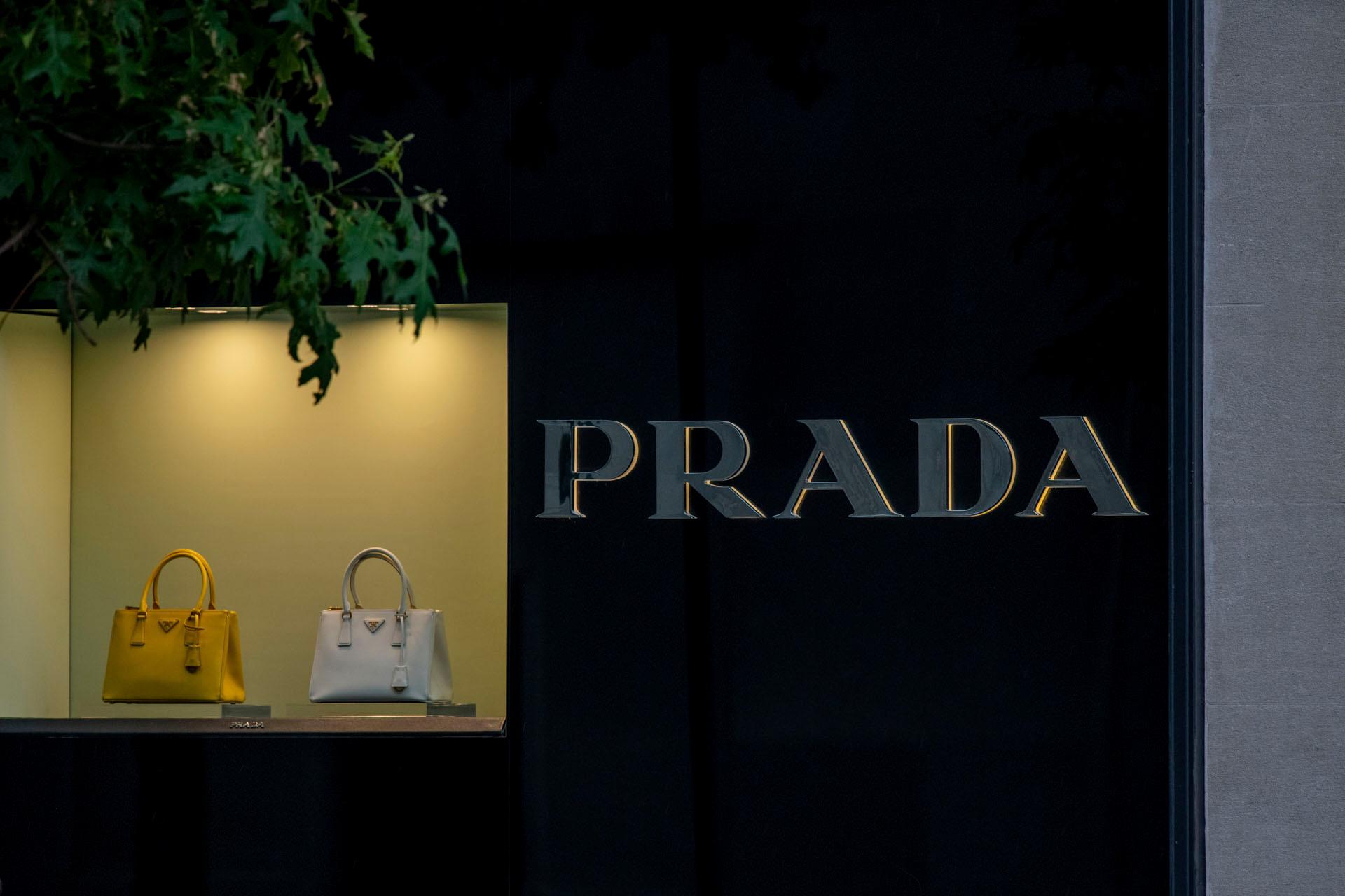 Storefront featuring the Prada logo and a display case with two handbags, one yellow and one white, under soft lighting. A tree partially shadows the scene.