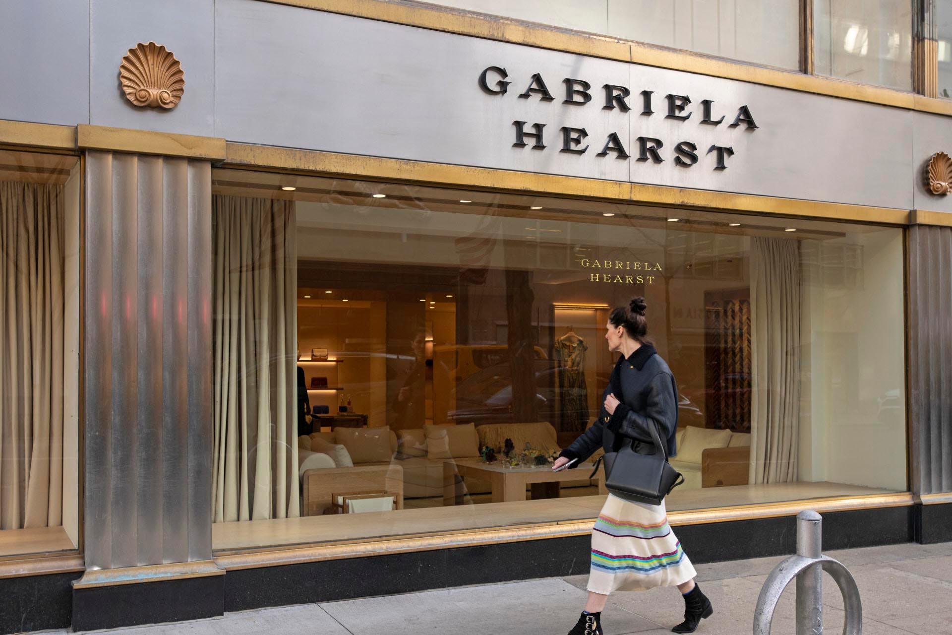A woman in a dark coat and striped skirt walks past the Gabriela Hearst storefront. The boutique's window displays elegant seating and decor.