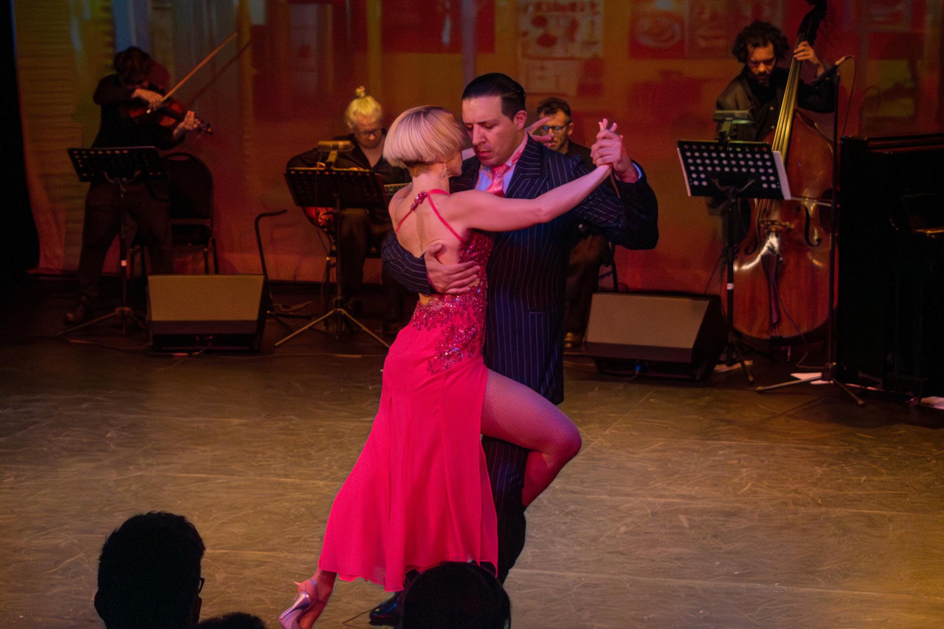 A couple dances tango passionately on stage; the woman wears a red dress and heels, the man a dark pinstripe suit. Musicians playing string instruments and double bass perform in the background.