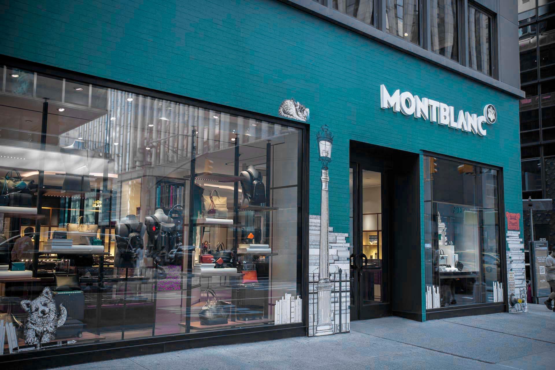 Street view of a Montblanc storefront with large windows displaying luxury items. The exterior is teal with white lettering. Stylized illustrations of books and cityscapes decorate the windows. The interior is visible, showcasing pens and leather goods.