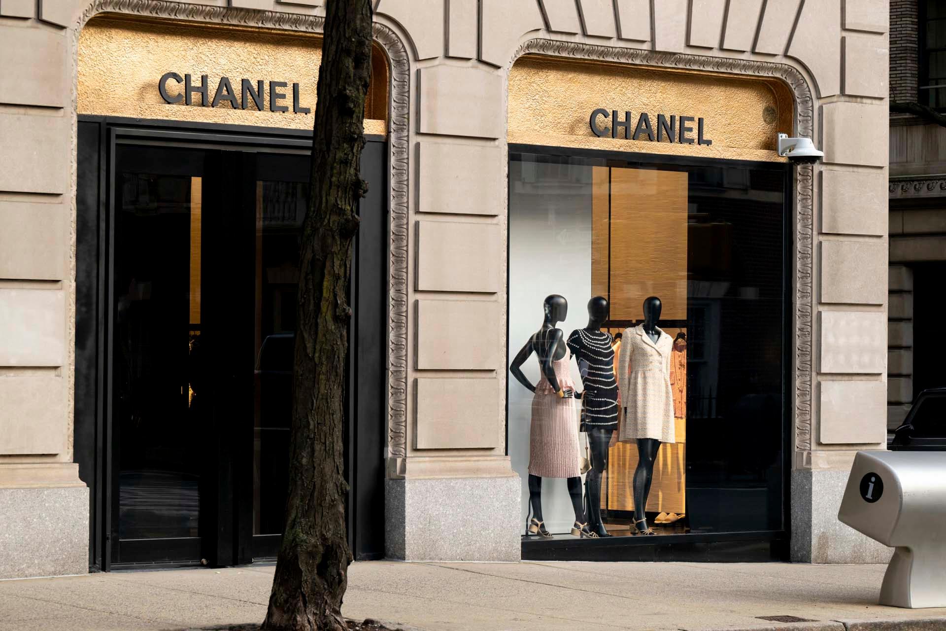 Street view of a Chanel store with two large black-framed windows. A tree partially obscures the entrance. Three mannequins in chic outfits are displayed in the other window. "CHANEL" signs are above the windows on gold backgrounds.