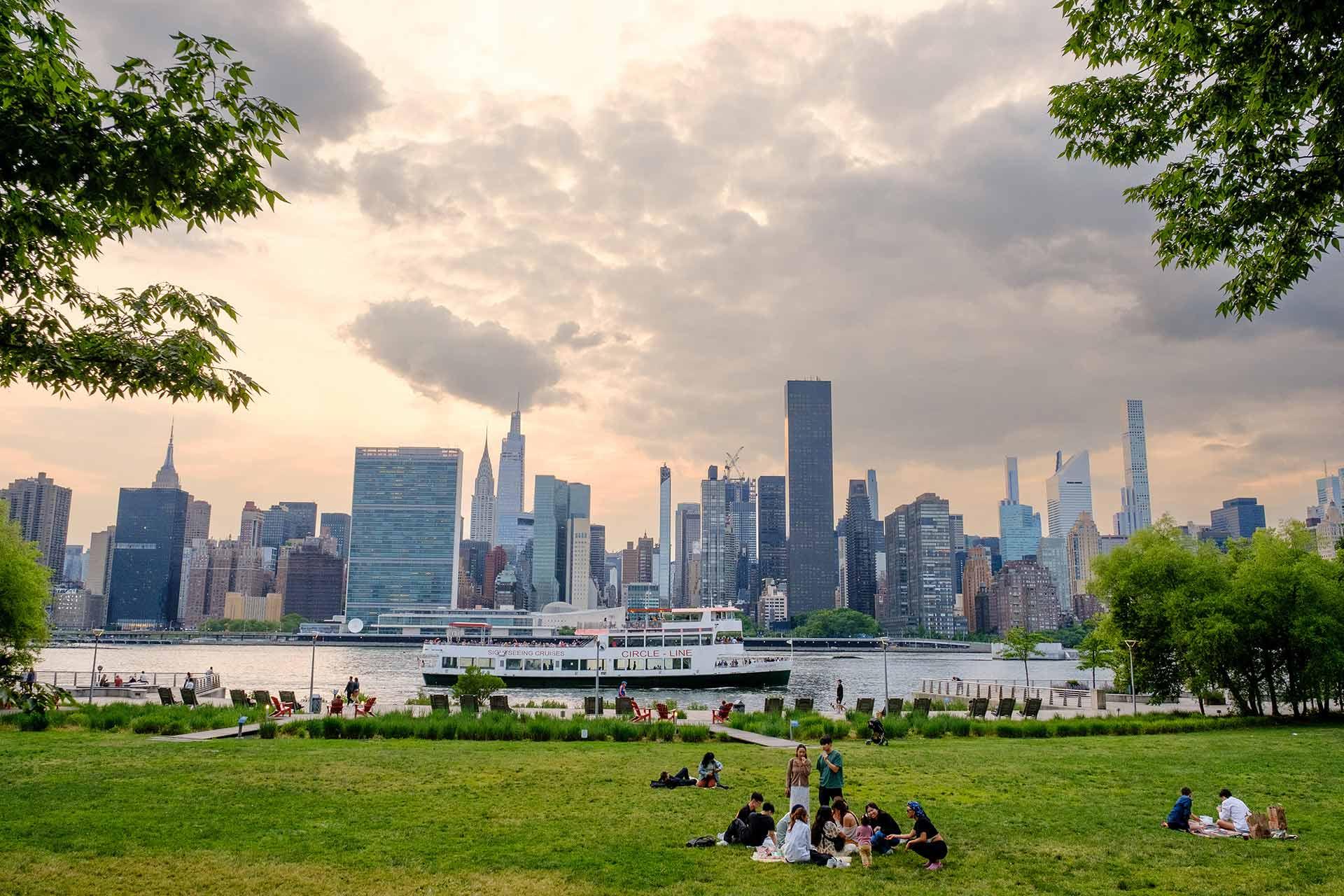 People relax on a grassy park overlooking the East River, with the New York City skyline and skyscrapers in the background under a cloudy sky. Trees frame the scene, and a boat is visible on the river.