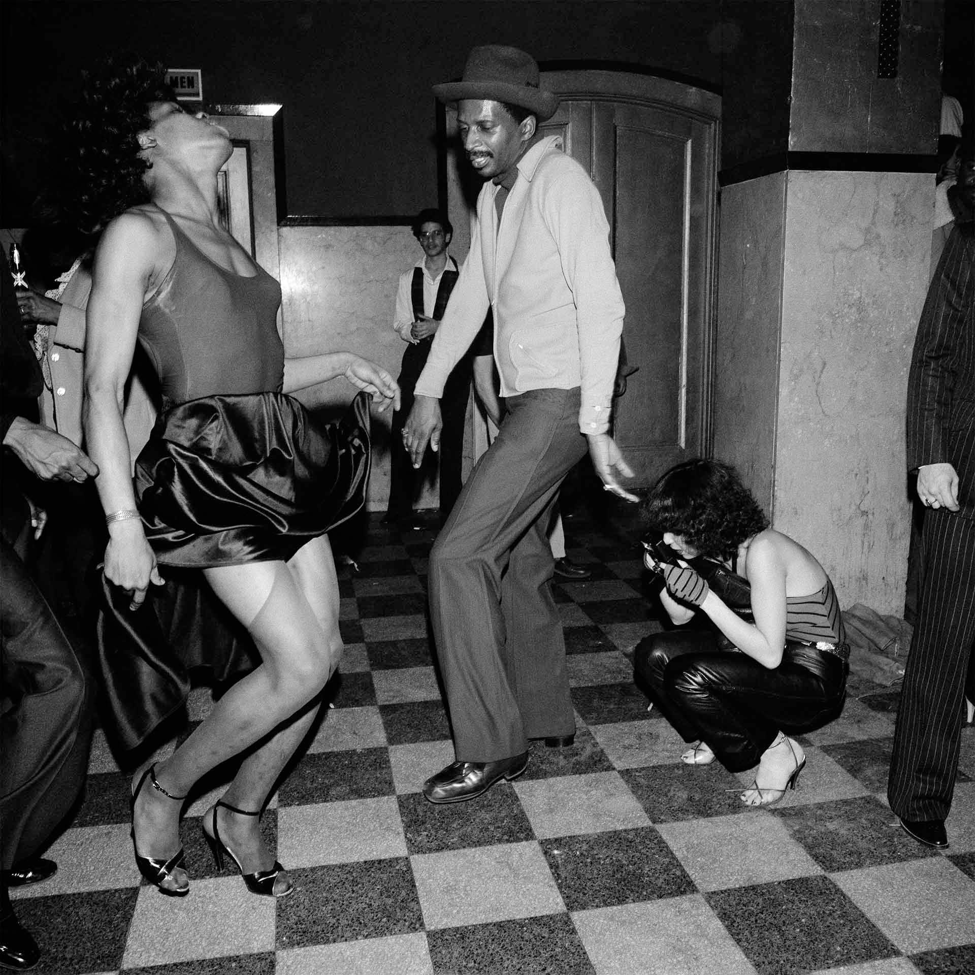 Two people dance energetically on a checkered floor at a nightclub while a photographer crouches nearby, capturing the moment. The scene is lively and joyful, set indoors with dim lighting.